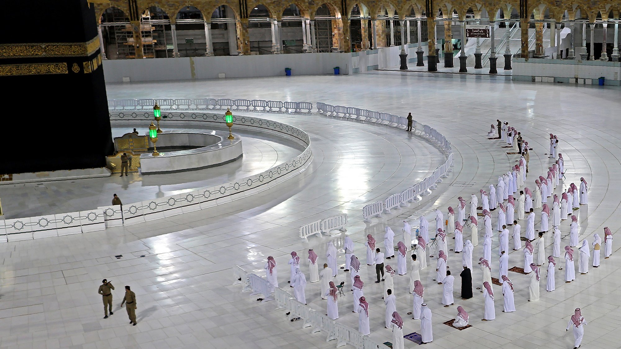 L'image montre une scène à la Kaaba, située à La Mecque. Plusieurs personnes en vêtements traditionnels blancs, souvent appelés " Ihram", sont disposées en rangées, apparemment en train de prier. On peut voir la Kaaba, un cube noir avec des détails dorés, au centre du sanctuaire. Des barrières de sécurité sont présentes pour délimiter les zones, et l'espace est vaste et lumineux, avec des éléments architecturaux en arrière-plan. Cette image évoque un moment de recueillement et de spiritualité dans un lieu sacré pour les musulmans.