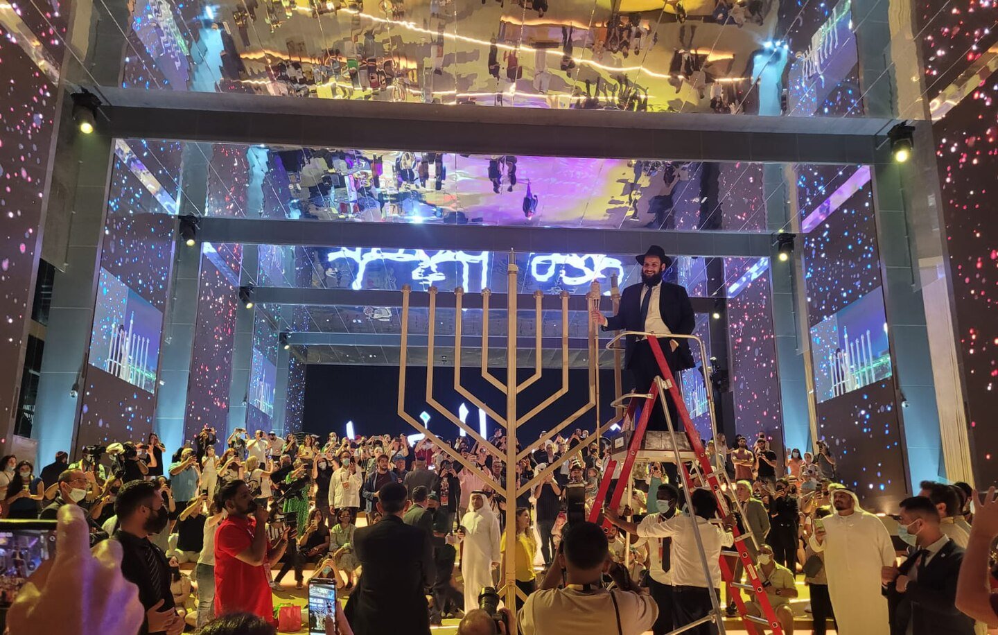 L'immagine mostra un evento festivo e affollato in un grande spazio, con una decorazione vibrante e luci colorate. Al centro, c'è una grande menorah, simbolo della tradizione ebraica, e una persona in cima a una scala che sembra essere coinvolta nella cerimonia di accensione. Il pubblico è composto da diverse persone in abiti tradizionali e moderni, tutte rivolte verso il palco. Il soffitto è riflettente, amplificando l'atmosfera festiva, mentre sullo sfondo si possono notare schermi luminosi e decorazioni artistiche. L'intera scena trasmette un senso di celebrazione e comunità.