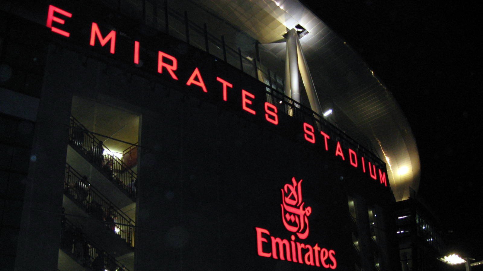 L'image montre l'extérieur de l'Emirates Stadium, qui est la maison du club de football Arsenal, à Londres. Le bâtiment est illuminé, avec le nom "EMIRATES STADIUM" en lettres rouges lumineuses bien visibles. On peut également voir le logo d'Emirates, une compagnie aérienne, mis en évidence. L'ambiance semble animée, suggérant qu'il y a probablement un événement ou un match en cours, avec des personnes rassemblées autour de la façade du stade.