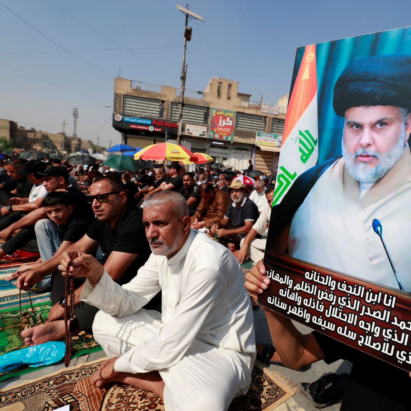 L'image montre une manifestation ou une assemblée publique. De nombreuses personnes sont assises sur des tapis, probablement pour un événement religieux ou politique. Un homme tient une grande affiche avec une photo d'un leader charismatique, visiblement respecté par les participants. Les drapeaux irakiens et d'autres décorations sont présents, ajoutant une ambiance colorée à la scène. Les participants semblent engagés dans une activité collective, reflétant un fort sentiment de communauté et de solidarité.