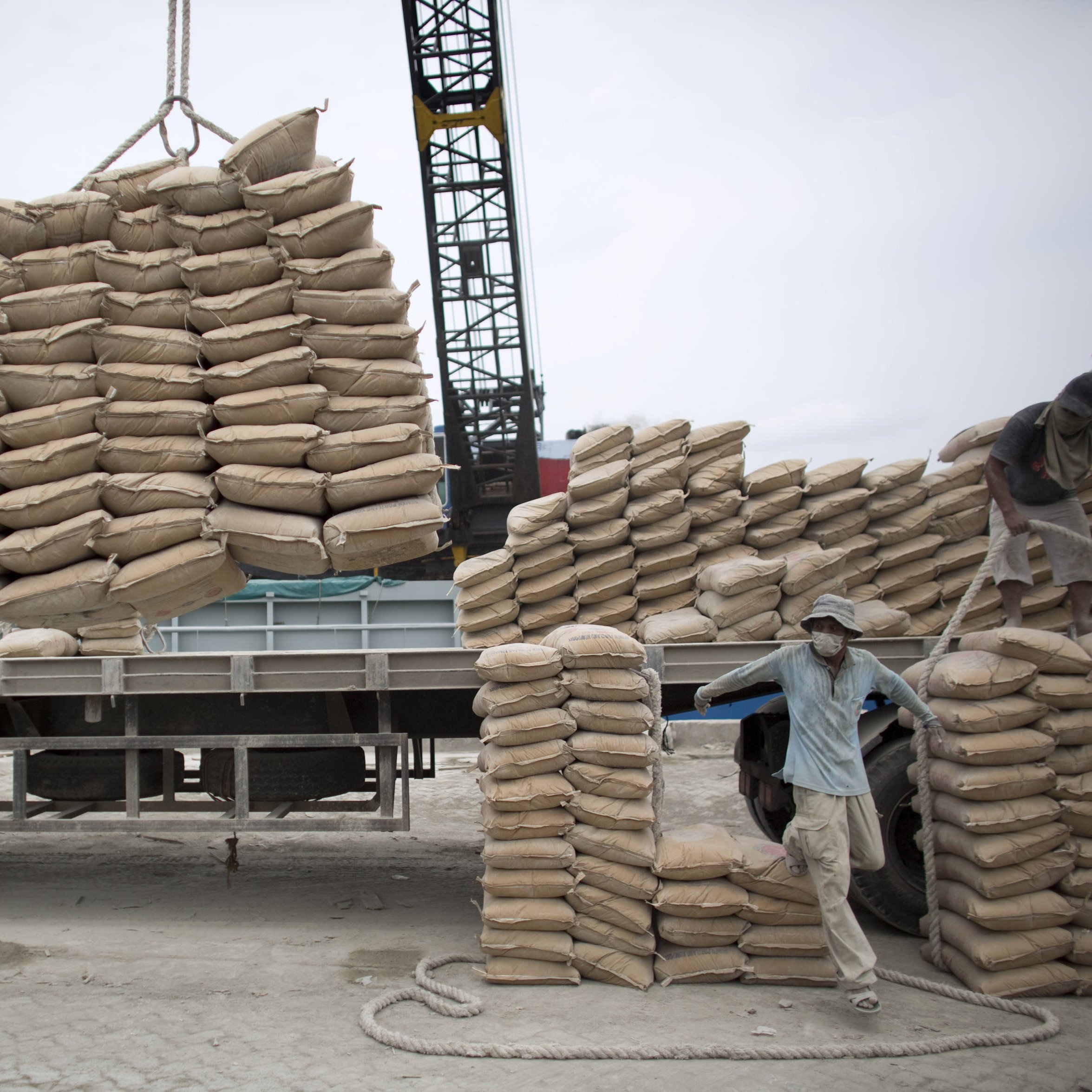 تظهر الصورة عمالًا يقومون بتحميل أكياس من مواد معينة على شاحنة. يبدو أن الأكياس مكدسة بعناية على الشاحنة، حيث يتم رفع بعضها باستخدام رافعة. يتركز العمل على تنظيم الأكياس وضمان تحميلها بشكل صحيح لتسهيل النقل. السماء تبدو غائمة، مما يوحي بأن العمل يتم في يوم غير مشمس.