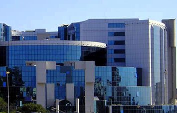 L'image montre un bâtiment moderne au design architectural contemporain. La façade est principalement en verre, ce qui reflète la lumière et les bâtiments environnants. Des formes géométriques et des courbes sont bien mises en valeur, et on peut distinguer des éléments en métal. Le paysage autour est agrémenté d'arbres et de plantes, suggérant que le bâtiment est situé dans une zone urbaine. L'ensemble dégage une impression de sophistication et de modernité.