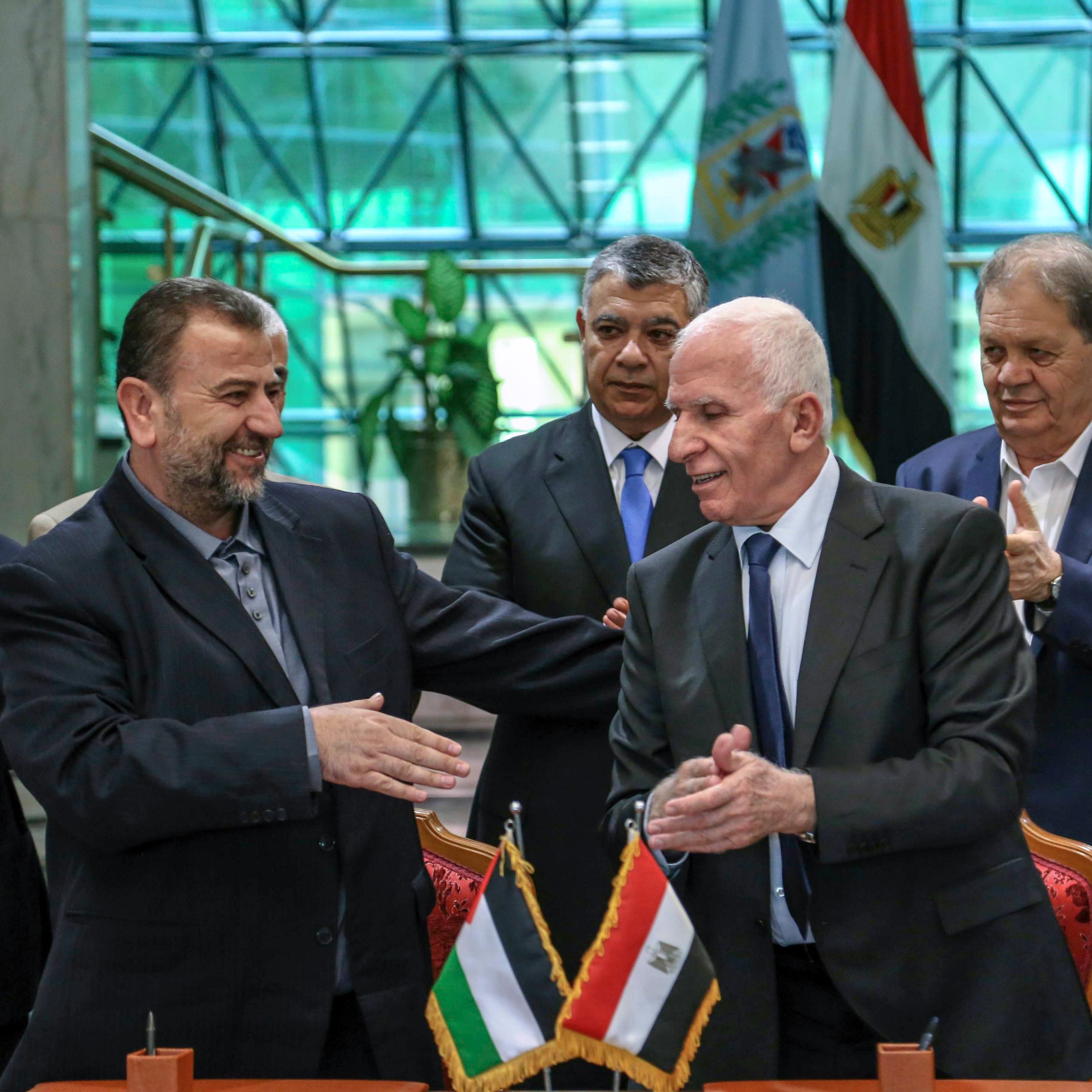 L'image montre un groupe de personnes lors d'une cérémonie officielle. À l'avant, deux hommes se serrent la main avec des sourires, suggérant un accord ou un partenariat. Ils sont entourés d'autres individus qui applaudissent, indiquant un moment de célébration. À l'arrière-plan, on peut distinguer des drapeaux, probablement ceux de la Palestine et de l'Égypte, ce qui laisse penser que cette rencontre est d'une importance politique. L'environnement semble être un bâtiment officiel, avec de grandes fenêtres laissant entrer la lumière.