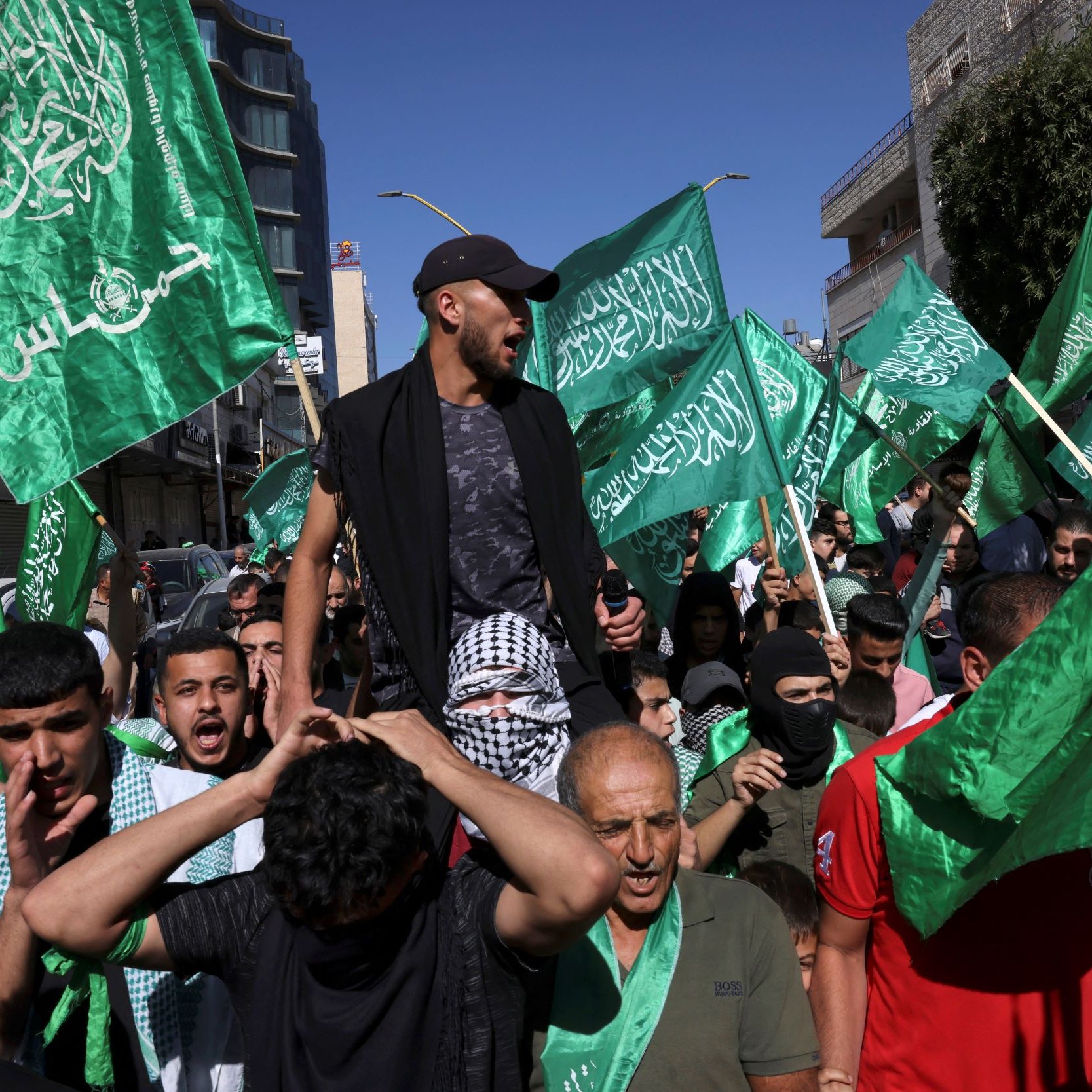 L'immagine mostra un gruppo di persone in una manifestazione. Molti partecipanti brandiscono bandiere verdi, che possono simboleggiare un movimento politico o religioso. Alcuni individui sembrano esprimere forti emozioni, con sorrisi e grida, mentre uno di loro è in piedi sopra la folla, probabilmente cercando di guidare o incoraggiare i manifestanti. Lo sfondo presenta edifici e un cielo sereno, suggerendo che l'evento si svolge all'aperto in una città.