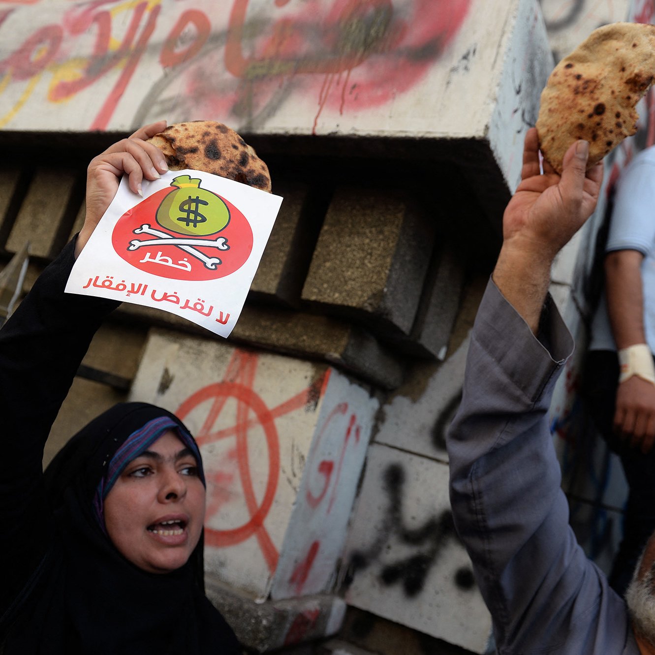 الصورة تظهر مجموعة من الأشخاص في تظاهرة. في المقدمة، يوجد رجل وامرأة يحملان لافتات. المرأة ترتدي الحجاب وتبدي حماسًا بالرفع لافتة مكتوب عليها شعار، بينما الرجل يحمل قطعة خبز. في الخلفية، تظهر جدران مكتوبة عليها شعارات وشعارات فنية. يبدو أن المتظاهرين يعبرون عن رأيهم حول موضوع معين، وملابسهم تشير إلى أجواء الاحتجاج.