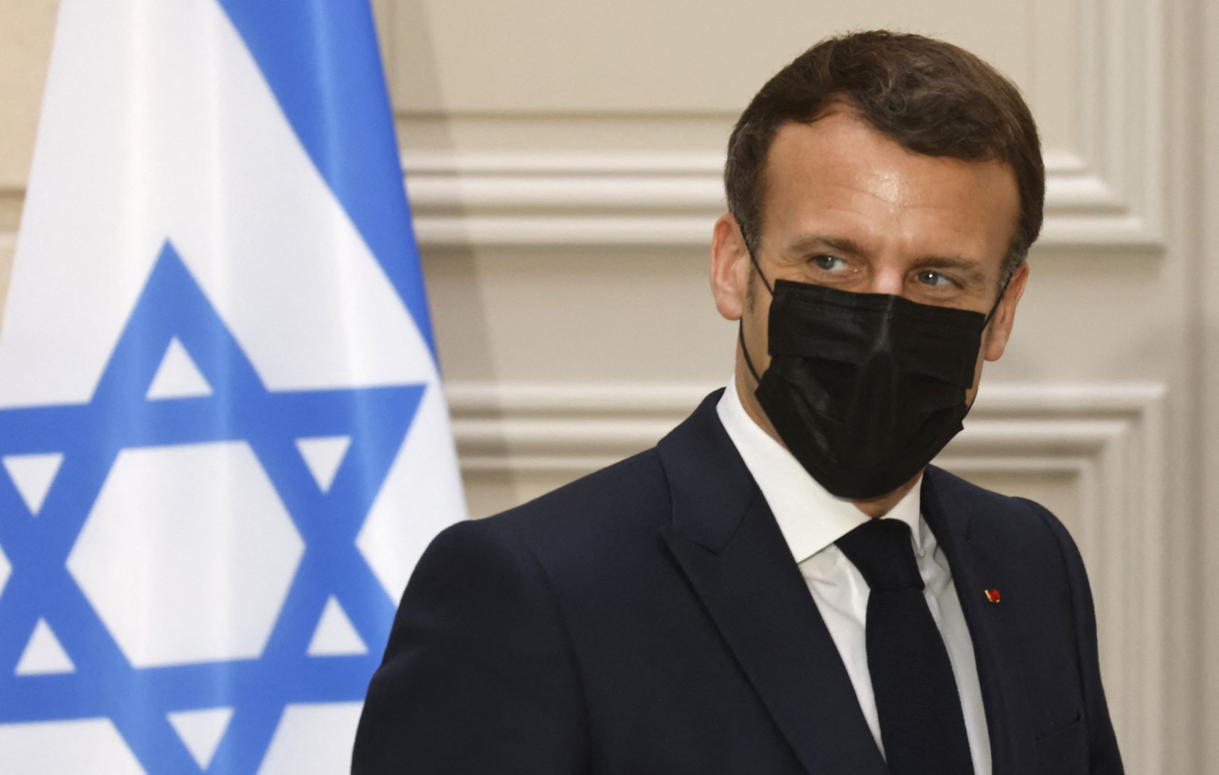 L'image montre une personne en costume, portant un masque noir, se tenant près d'un drapeau qui comporte une étoile de David. L'arrière-plan semble être un intérieur formel, peut-être un bureau ou une salle de réunion. La personne a une expression attentive et semble engagée dans une conversation.