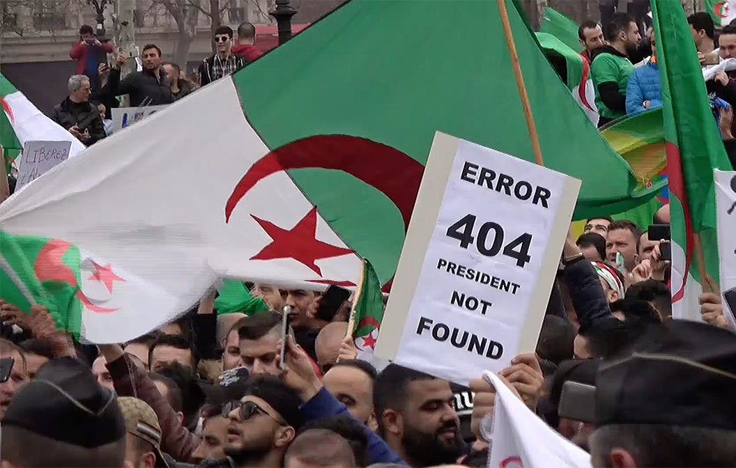 الصورة تظهر مجموعة من المحتجين في تظاهرة، يحملون أعلام الجزائر بالإضافة إلى لافتة مكتوب عليها "ERROR 404 PRESIDENT NOT FOUND". تعكس هذه الصورة مشاعر الاحتجاج والمطالبة بالتغيير السياسي، حيث يُظهر المتظاهرون تصميمهم على المطالبة بحقوقهم وبإصلاحات في البلاد. الأجواء تبدو حماسية ومليئة بالروح الوطنية.
