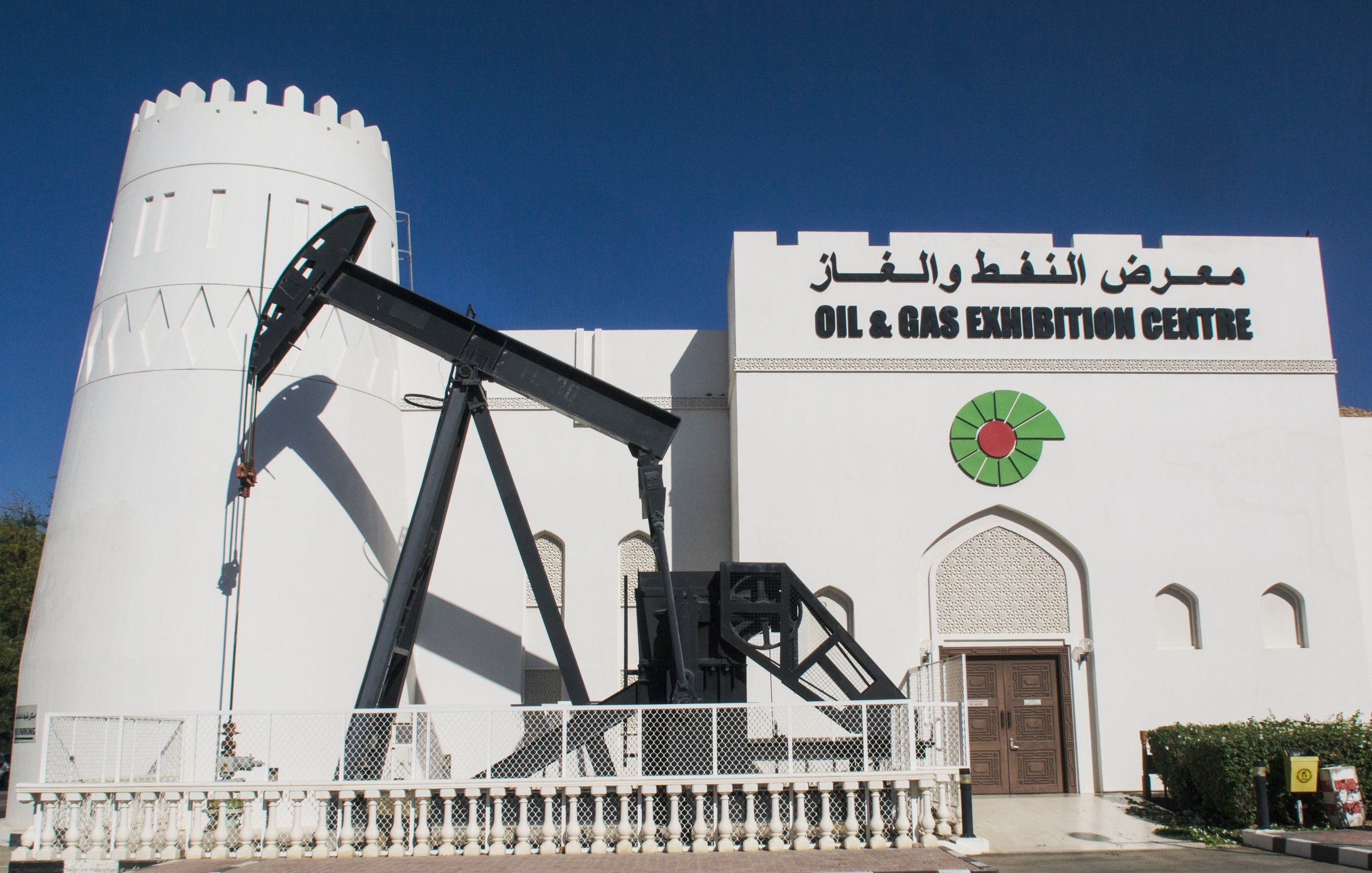 The image features a white, castle-like building with a prominent sign that reads "OIL & GAS EXHIBITION CENTRE" in both English and Arabic. To the left of the entrance, there is an oil pump jack, a piece of machinery commonly used in oil extraction. The building has a decorative design with a combination of traditional and modern architectural elements. The sky is clear and blue, creating a vibrant backdrop.