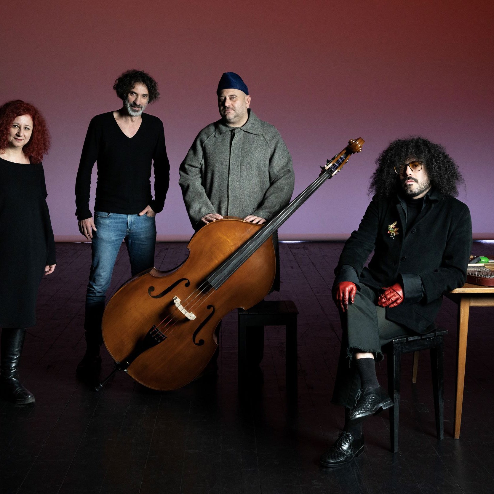 L'image présente un groupe de quatre personnes posant ensemble sur scène. Au centre, il y a un grand instrument de musique, probablement un instrument à cordes, qui attire l'attention. À gauche, une femme avec des cheveux roux bouclés se tient avec une expression sérieuse. À côté, un homme avec des cheveux frisés et un pull noir semble également concentré. Au centre, près de l'instrument, un homme porte une tenue grise et un couvre-chef. Enfin, à droite, un autre homme avec des lunettes et des gants rouges est assis à une table avec divers objets, ayant l'air intrigant. L'éclairage de la scène crée une ambiance artistique et captivante.