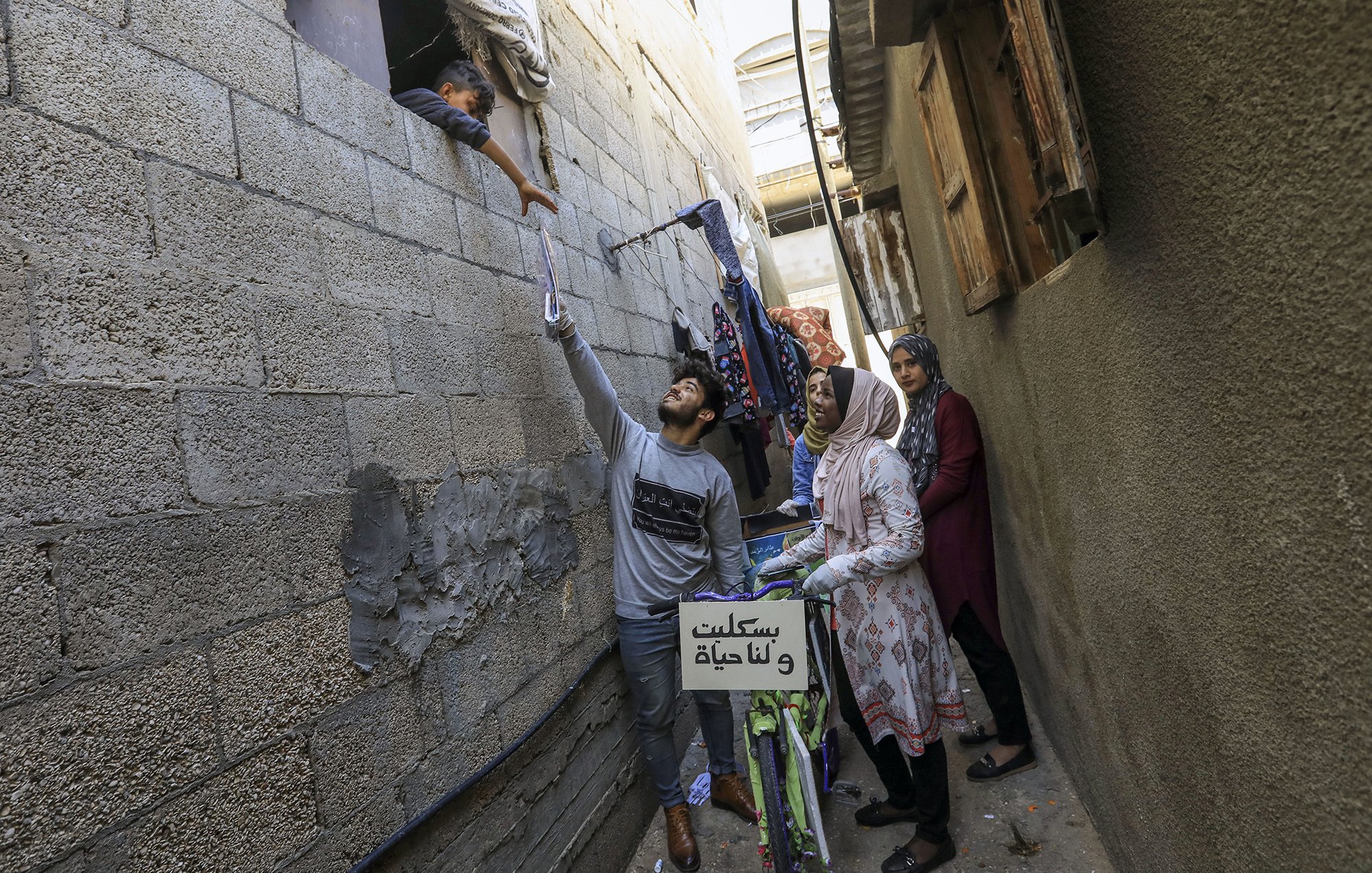L'image montre une scène dans une ruelle étroite, probablement dans un quartier urbain. On y voit un jeune homme qui tend la main vers une fenêtre, où un enfant semble interagir avec lui. À l'arrière-plan, une femme, probablement la mère de l'enfant, observe la scène. Une autre femme se tient à côté, regardant également. Des vêtements sont suspendus à l'extérieur, ajoutant une touche de vie quotidienne à l'image. Une bicyclette est visible, avec un panneau sur le guidon. L'atmosphère semble chaleureuse et familiale, malgré le contexte urbain.