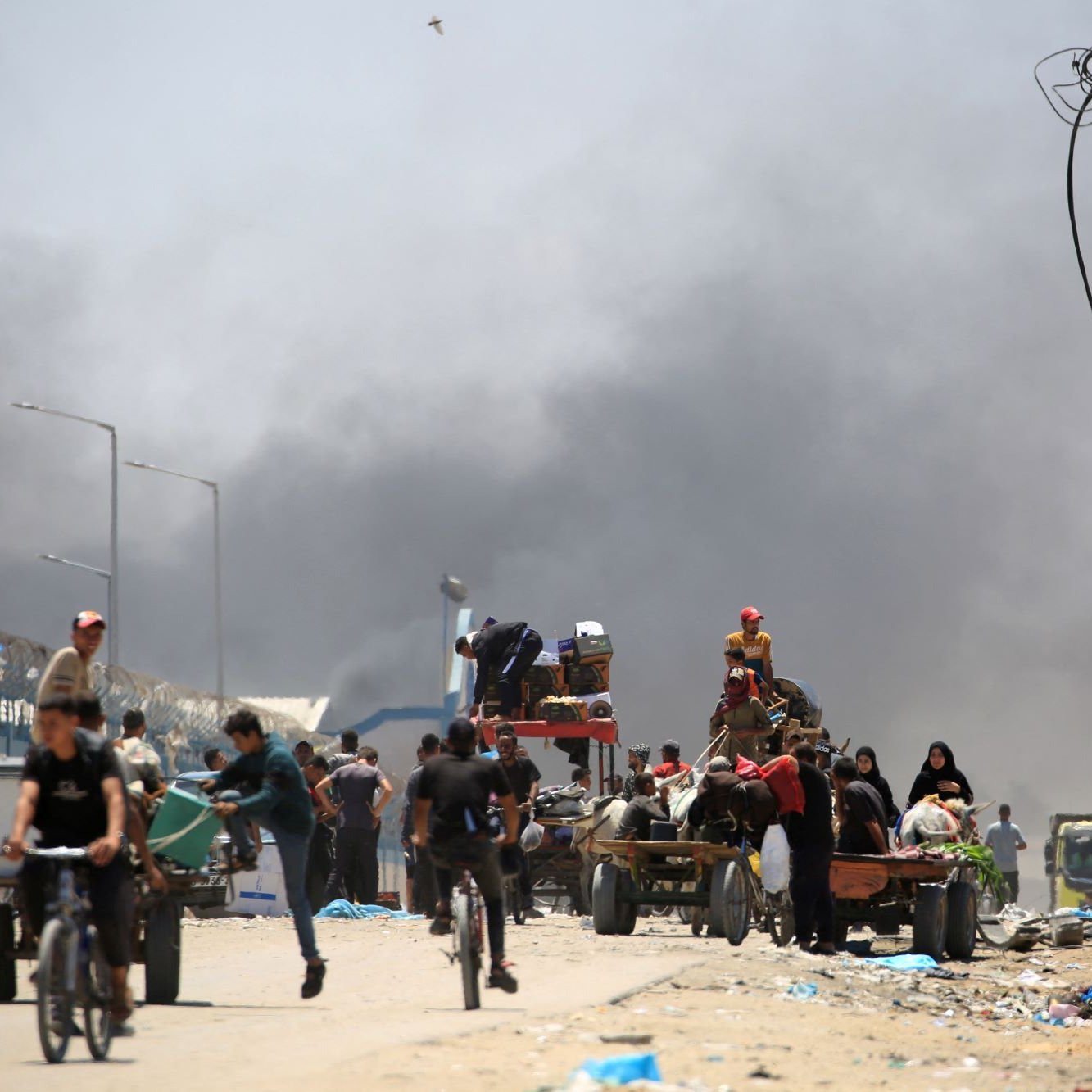 L'image montre une scène chaotique dans un environnement urbain. On peut voir des gens marchant et roulant à vélo le long d'une route, tandis qu'un groupe semble être occupé à charger ou décharger des marchandises sur des chariots. En arrière-plan, de la fumée s'élève, suggérant qu'un incendie ou une explosion a eu lieu. L'ambiance semble tendue et désordonnée, avec des câbles électriques visibles et des structures urbaines en arrière-plan.