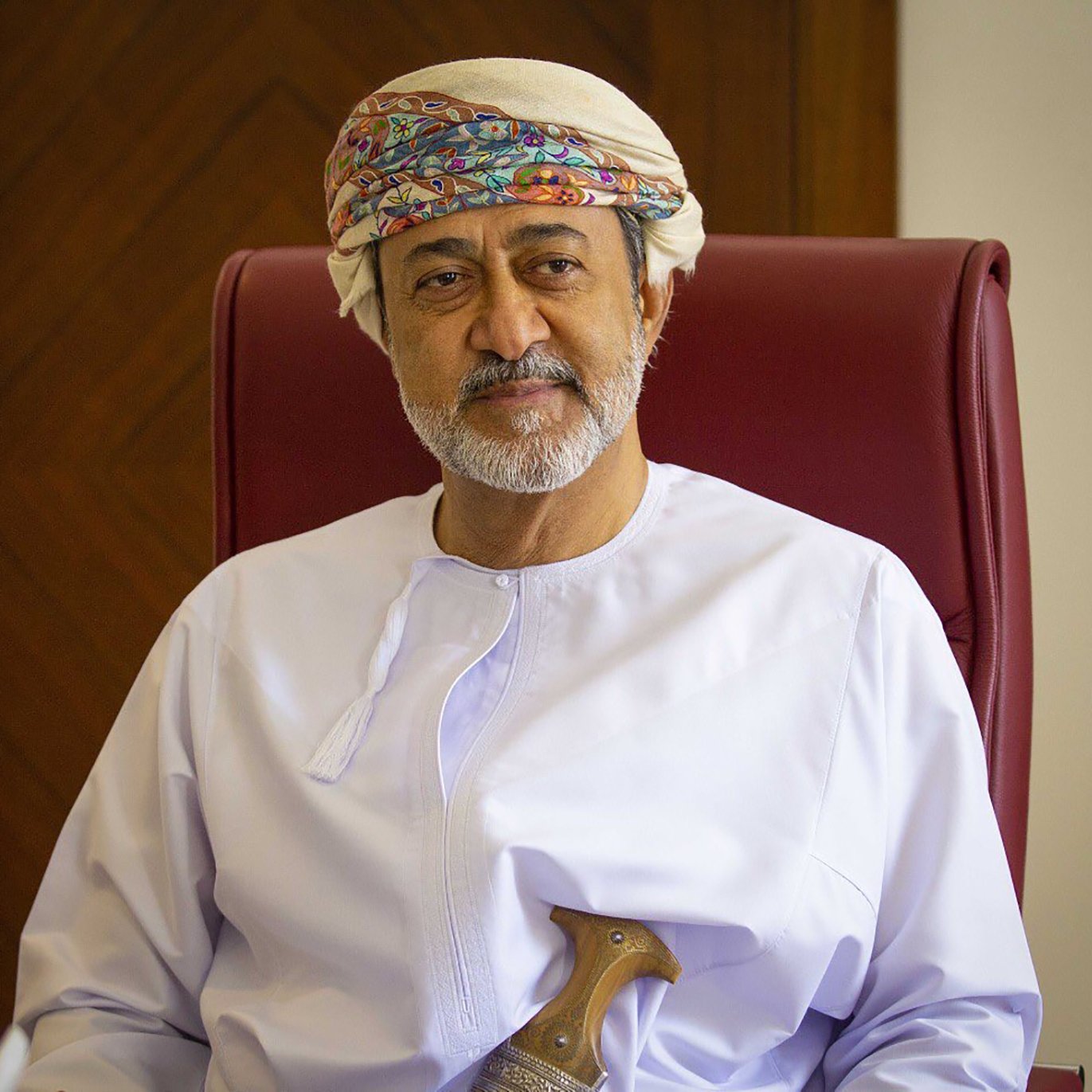 الصورة تظهر شخصًا يجلس على كرسي. يرتدي الملابس التقليدية العمانية، حيث يظهر بجلابية بيضاء وعصابة رأس مزخرفة. في يده يوجد خنجر عماني تقليدي. خلفه خلفية خشبية بسيطة. يعكس تعبير الوجه الهدوء والثقة.