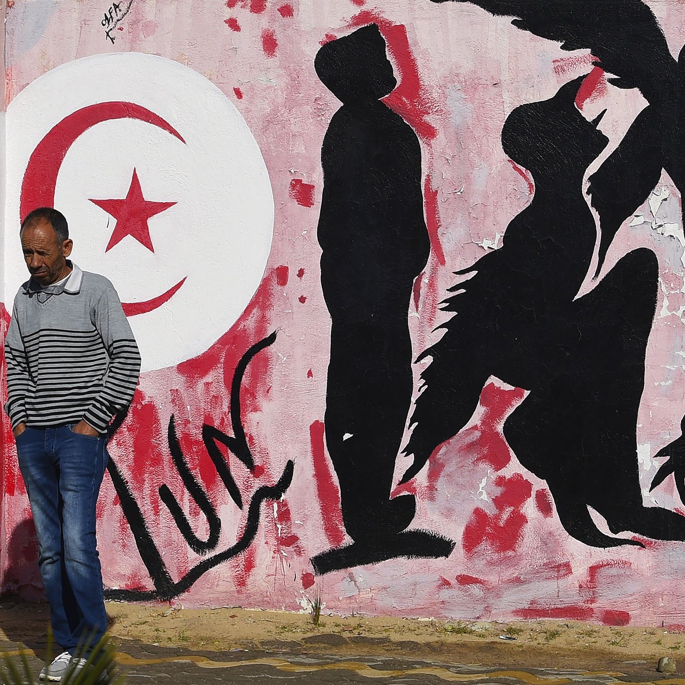 L'immagine mostra un murale su un muro bianco, caratterizzato da una palette di colori rossi e neri. Sullo sfondo, è presente un grande simbolo della luna crescente e una stella, simboli spesso associati alla bandiera tunisina. Al centro, ci sono delle silhouette di figure umane che appaiono in diverse posizioni: alcune sono in piedi, altre sembrano inginocchiarsi o muoversi in modo dinamico. In primo piano, un uomo con una maglietta a strisce passeggia accanto al murale, creando un contrasto tra l'arte e la vita quotidiana. L'atmosfera trasmette un senso di cultura e identità, evidentemente legato alla Tunisia.