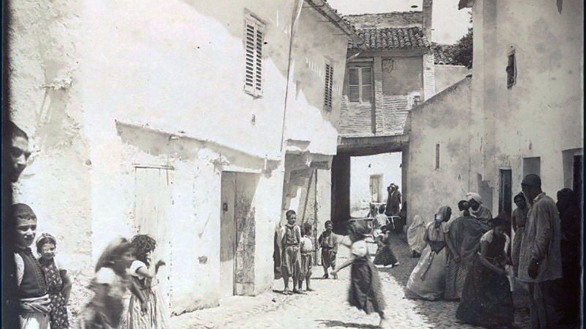 L'image représente une scène de rue dans un quartier de Constantine, en Algérie. On y voit des maisons blanches avec des volets, et une ruelle pavée qui semble étroite. Plusieurs personnes, y compris des enfants, sont visibles, certaines marchant et d'autres se tenant debout. Les vêtements des habitants semblent traditionnels, reflet d'une époque passée. L'atmosphère générale évoque une vie quotidienne animée dans un contexte historique.