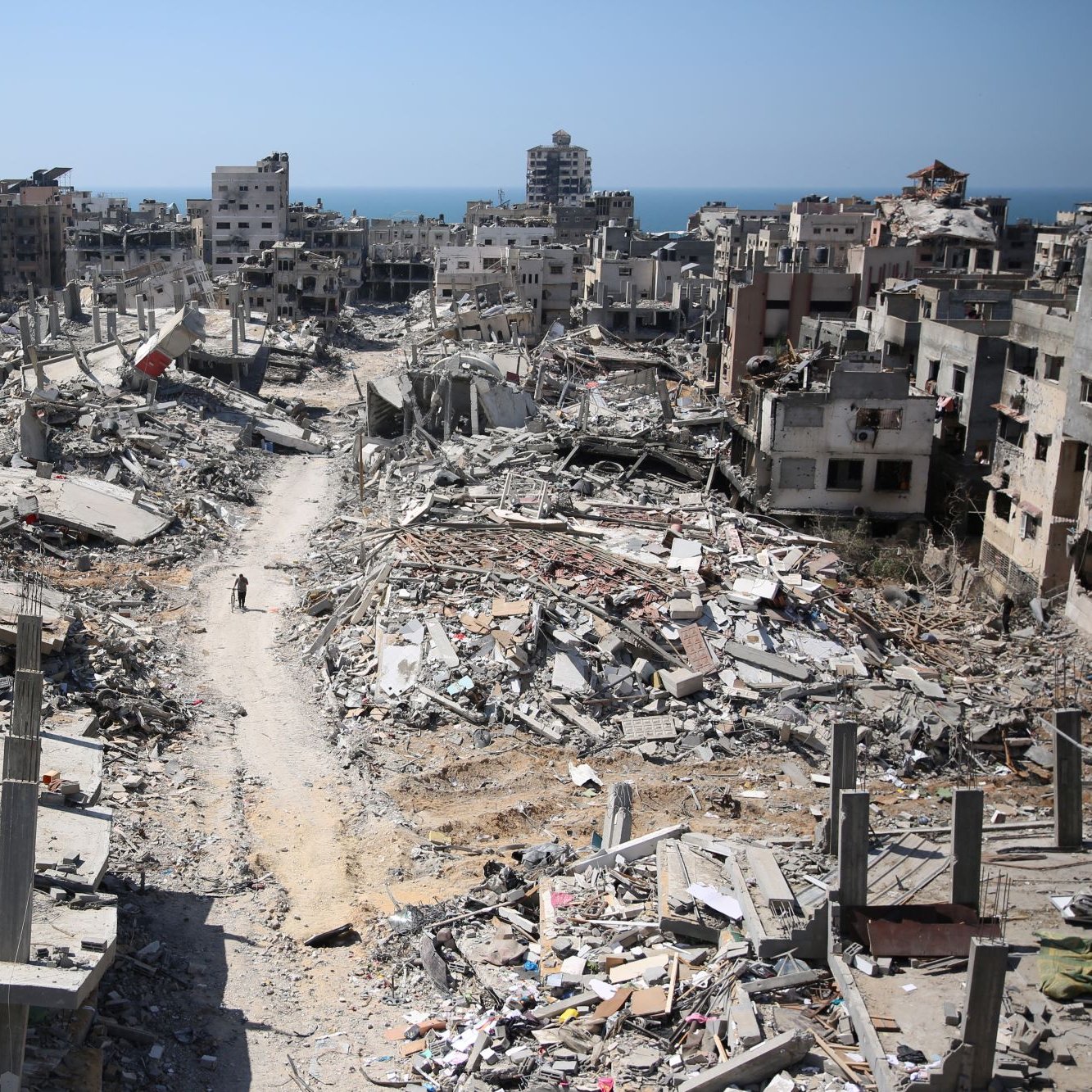 تصویر نشان‌دهنده یک منطقه ویران شده است که ساختمان‌های تخریب‌شده و خرابه‌ها در آن دیده می‌شوند. در پس‌زمینه، دریا نمایان است و آسمان آبی رنگ است. در این صحنه، یک مسیر خاکی به چشم می‌خورد که از میان ویرانه‌ها عبور می‌کند و در انتهای آن، فردی در حال حرکت است. این تصویر حس غم و ویرانی را منتقل می‌کند و نشان‌دهنده آسیب‌های ناشی از جنگ یا بلایای طبیعی است.