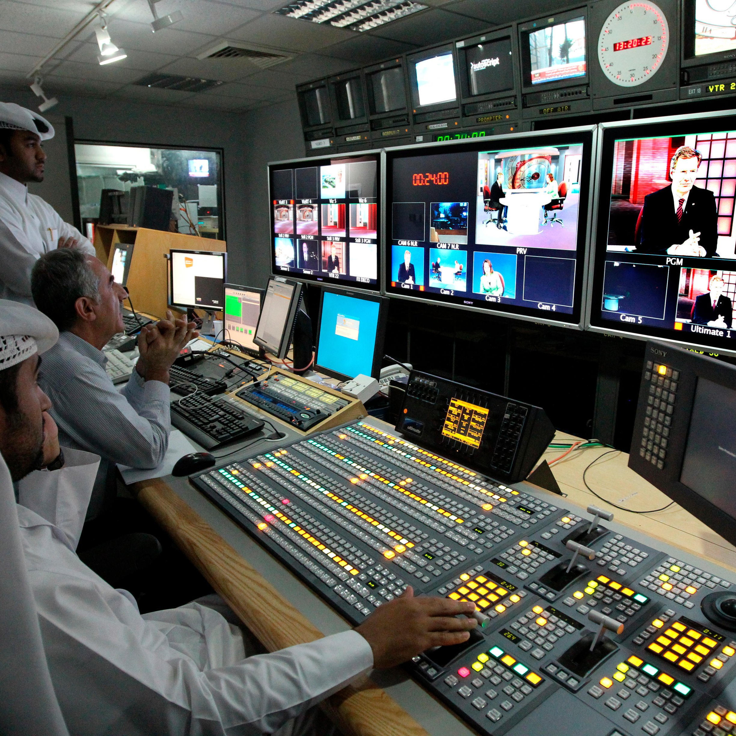 L'image montre un intérieur d'une salle de contrôle de diffusion. On y voit plusieurs personnes travaillant sur des panneaux de contrôle avec des écrans affichant différentes émissions. Au premier plan, une personne est assise devant une console, mesurant les niveaux et ajustant les paramètres. Deux autres personnes se tiennent derrière, observant attentivement les écrans. Les murs sont équipés de plusieurs moniteurs affichant des vidéos en direct ou des reportages. L'ambiance semble concentrée et professionnelle, typique d'un environnement de télévision.