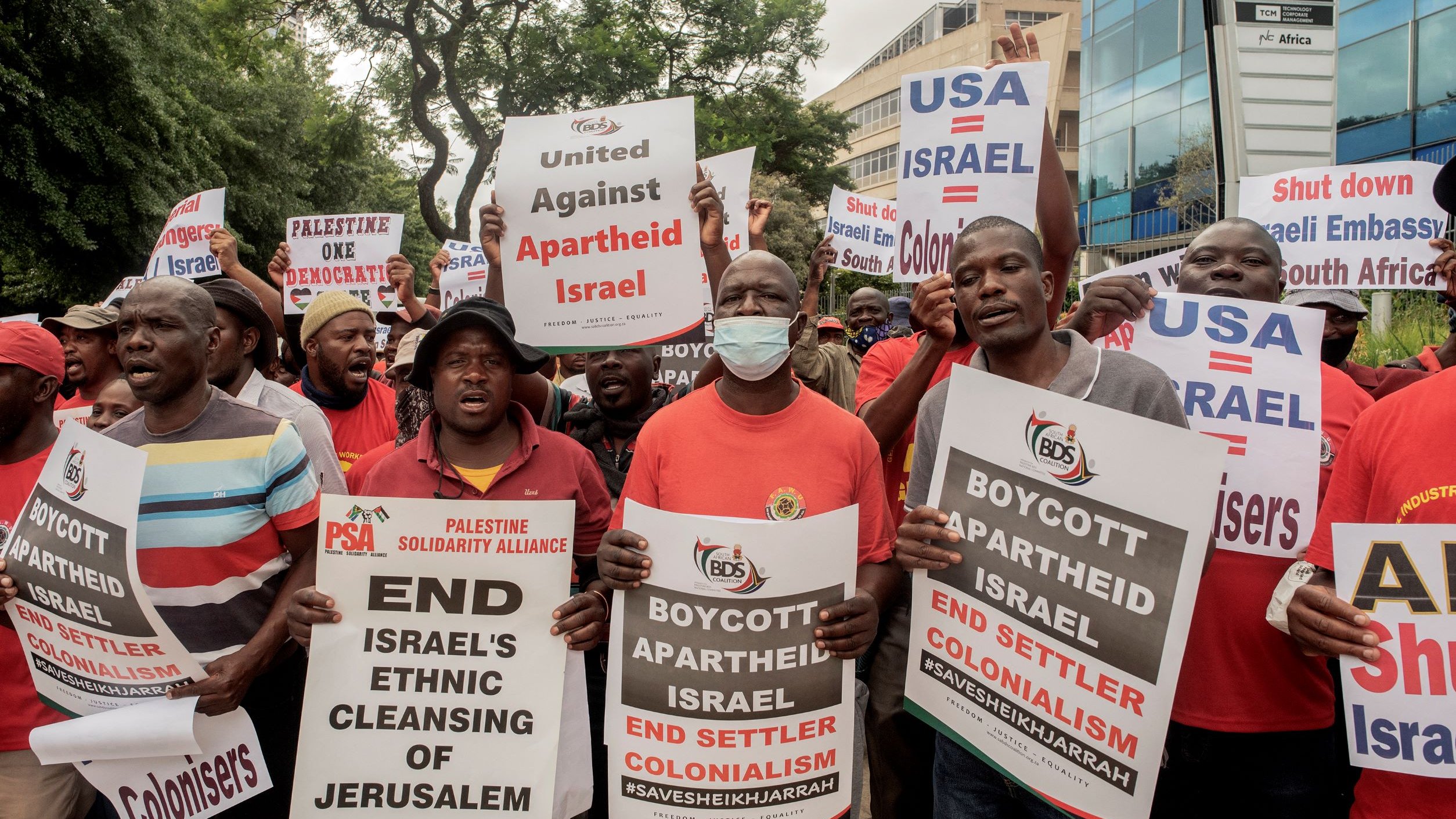 L'image montre une foule de manifestants tenant des pancartes et exprimant leur opposition à la politique israélienne, en particulier en ce qui concerne le traitement des Palestiniens. Les slogans sur les pancartes incluent des appels à mettre fin à l'apartheid et au colonialisme en Israël, ainsi qu'à boycotter certaines actions liées à ce pays. Les manifestants semblent très engagés dans leur cause, et l'atmosphère est dynamique et déterminée.