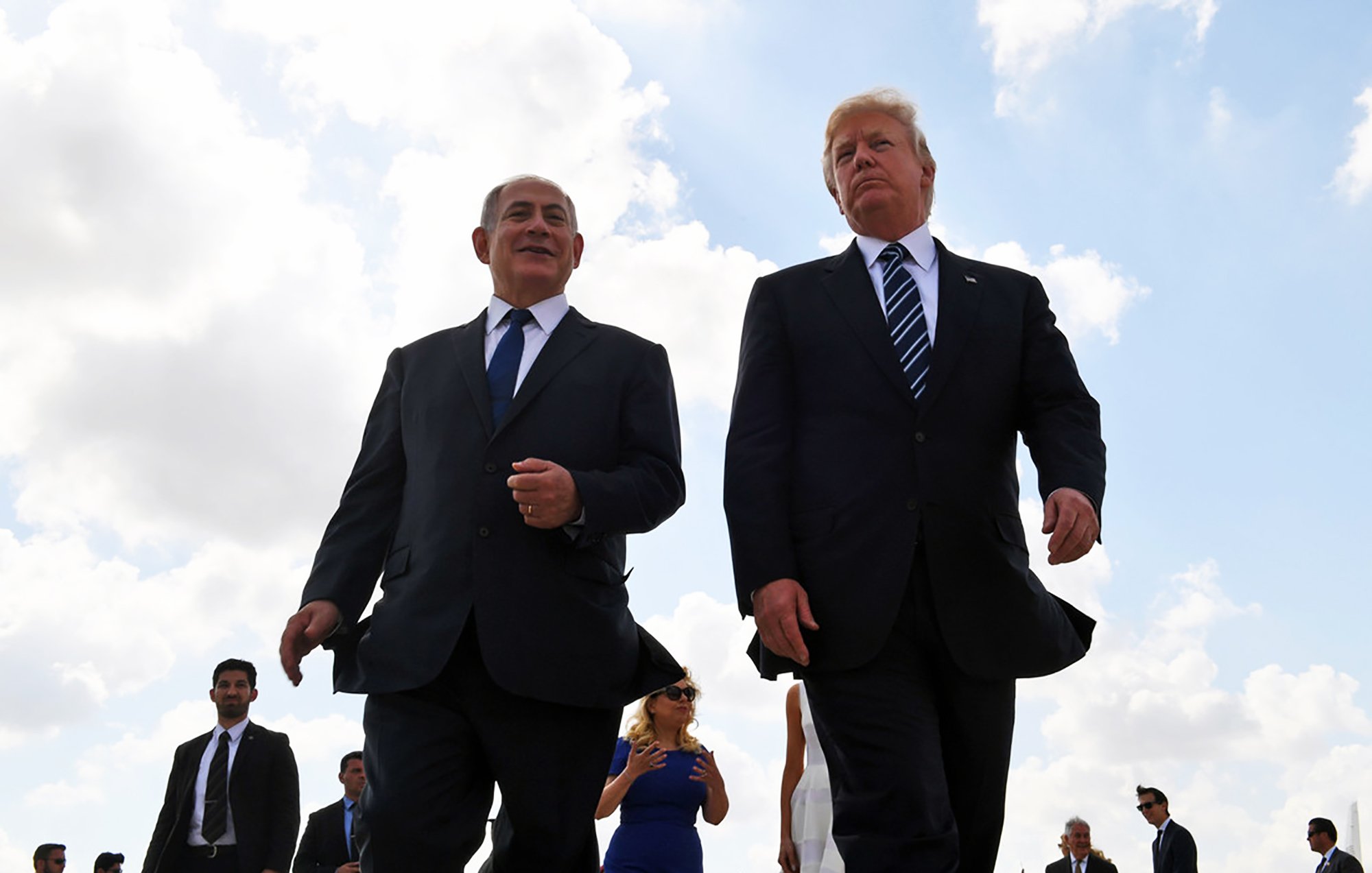 La imagen muestra a dos hombres caminando juntos al aire libre. Ambos visten trajes formales, con corbatas. Hay un grupo de personas detrás de ellos, y el cielo está parcialmente nublado. La escena transmite una sensación de formalidad y camaradería.