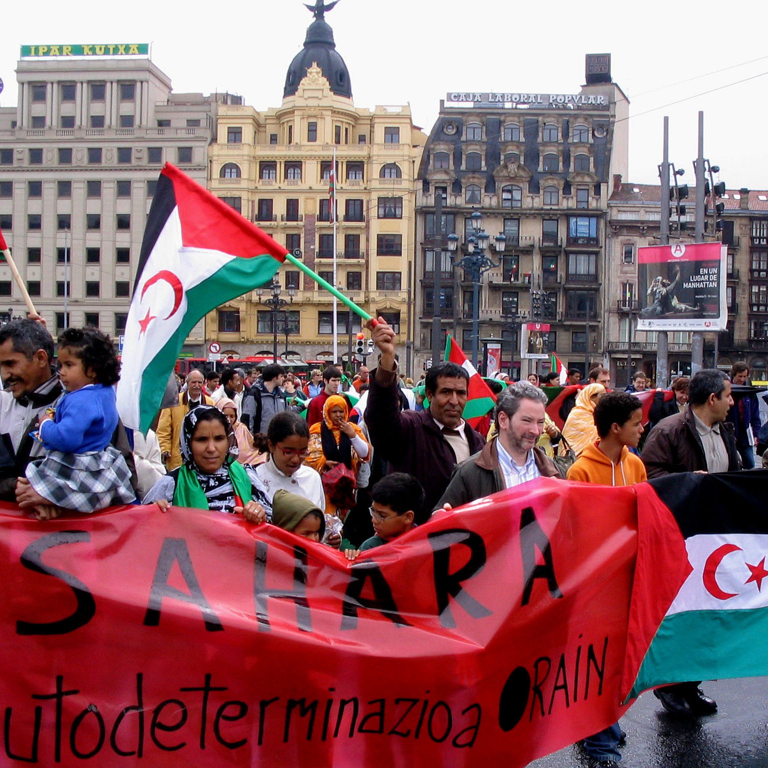 L'image montre une manifestation dans une ville, où un groupe de personnes tient des banderoles et des drapeaux, probablement en soutien à la cause du Sahara Occidental. Les participants, de différents âges, expriment leur solidarité avec des slogans affichés sur des pancartes en espagnol et en arabe. L'ambiance semble engagée et déterminée, mettant en avant un mouvement pour l'autodétermination. L'architecture autour d'eux indique que cela se déroule dans un espace urbain animé.