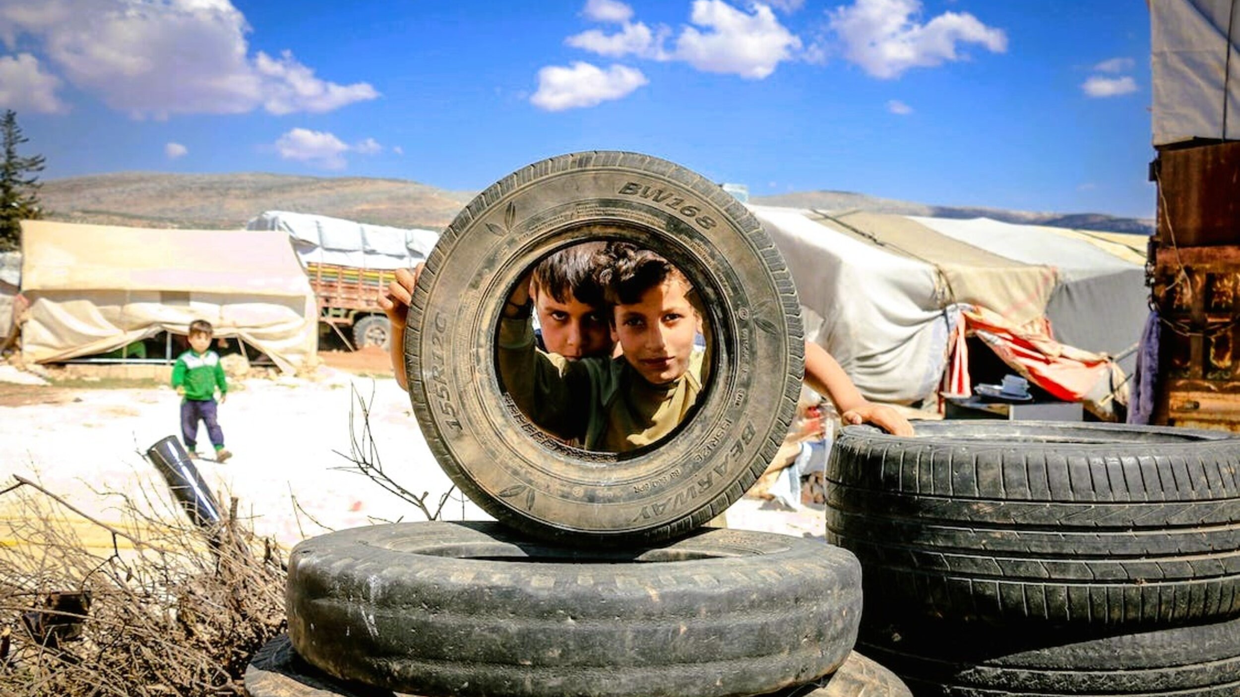 Nell'immagine si vedono due bambini che si affacciano attraverso un copertone di gomma, posto in orizzontale. I bambini, che sembrano curiosi e giocosi, sono circondati da altri copertoni impilati. Sullo sfondo si possono intravedere tende bianche e un paesaggio collinare sotto un cielo blu con alcune nuvole. L'atmosfera sembra essere di gioco e scoperta, nonostante l'ambiente circostante possa suggerire condizioni di vita difficili.