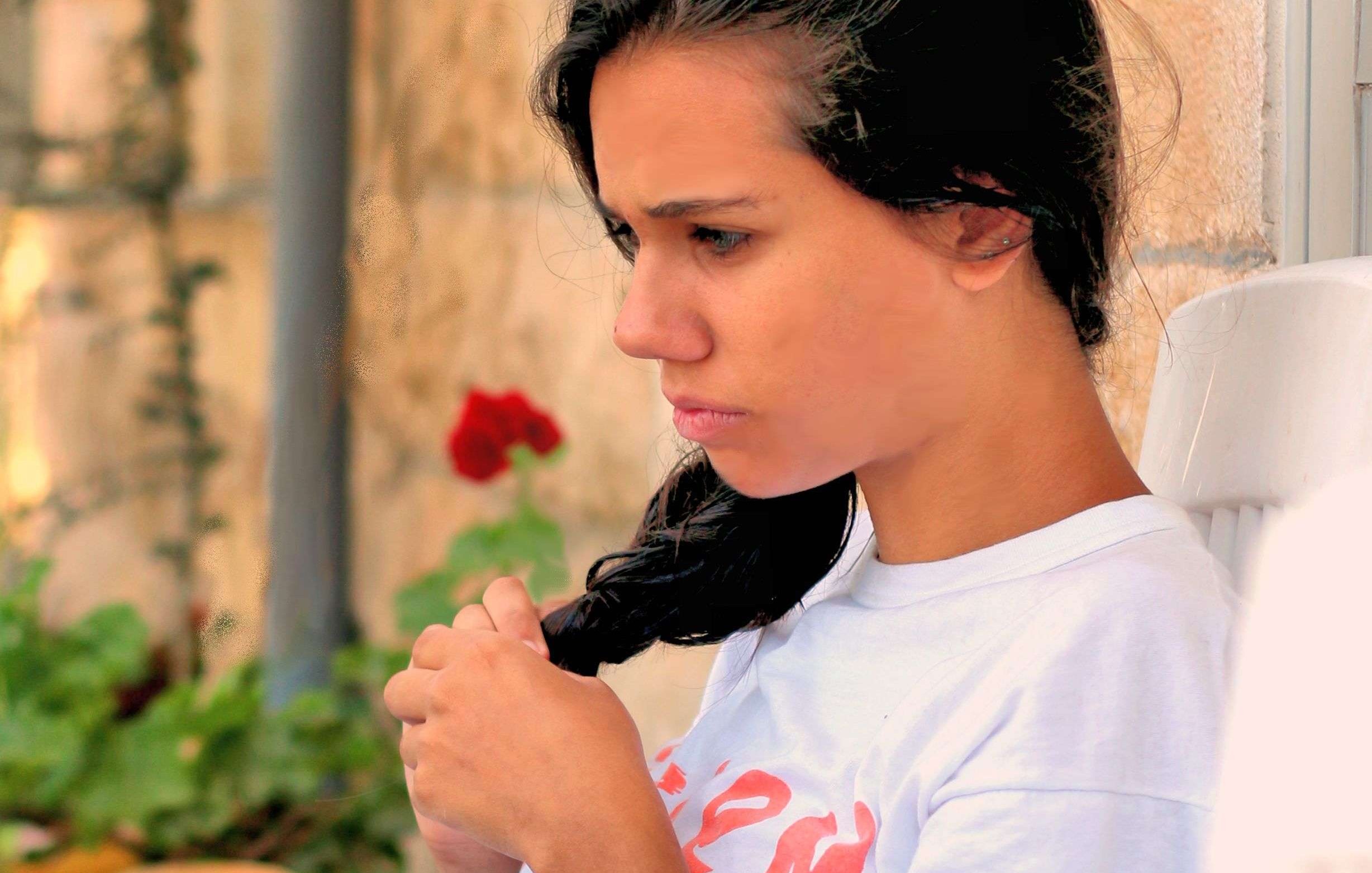 L'image montre une jeune femme assise sur une chaise, tournée légèrement sur le côté. Elle a les cheveux longs et noirs, et elle semble pensive, regardant au loin. Elle porte un t-shirt blanc avec un motif rouge. En arrière-plan, on aperçoit un mur en pierre et une plante verte dans un pot. L'atmosphère semble calme et introspective.