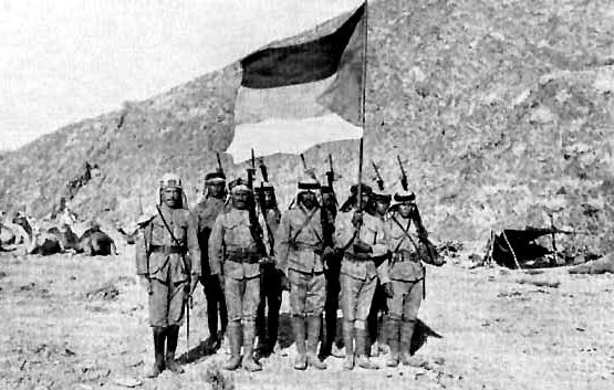 L'image représente un groupe de soldats posant devant un drapeau. Ils sont vêtus de uniforme militaire, portant des capes et des casques. En arrière-plan, on aperçoit un paysage montagneux et quelques tentes, suggérant un camp militaire. Les soldats, tenant leurs fusils, semblent être en position de parade ou de cérémonie. L'atmosphère de la photo évoque une période historique de conflit ou de guerre.