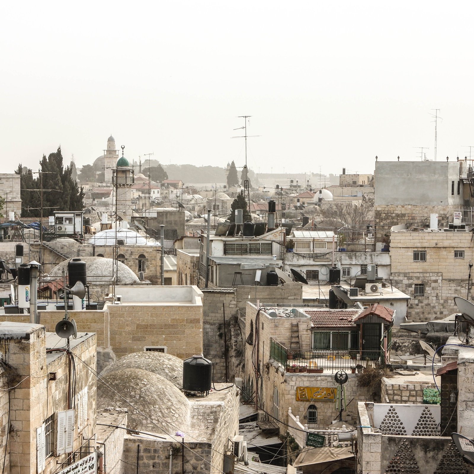 L'image montre un paysage urbain d'une ville ancienne, probablement Jérusalem. On peut voir une multitude de bâtiments en pierre avec des toits en tôle et des dômes, créant une texture variée. Les structures sont serrées les unes contre les autres, et quelques antennes de télévision sont visibles. Au loin, se distingue un minaret ou une coupole, indiquant la présence d'une mosquée. L'atmosphère semble un peu brumeuse, donnant une impression de chaleur et d'ancienneté à la scène.