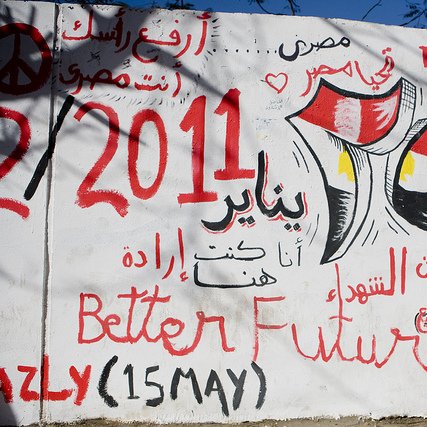 L'image montre un mur recouvert de graffitis en arabe et en anglais. Parmi les inscriptions, on trouve des références à l'Égypte, à la révolution de 2011, et des slogans tels que "Meilleur avenir" et des phrases évoquant la détermination et l'espoir. Les couleurs rouge, noir et jaune du drapeau égyptien sont visibles, renforçant le message patriotique et engagé. L'ensemble évoque une période de changement et de revendications sociales.