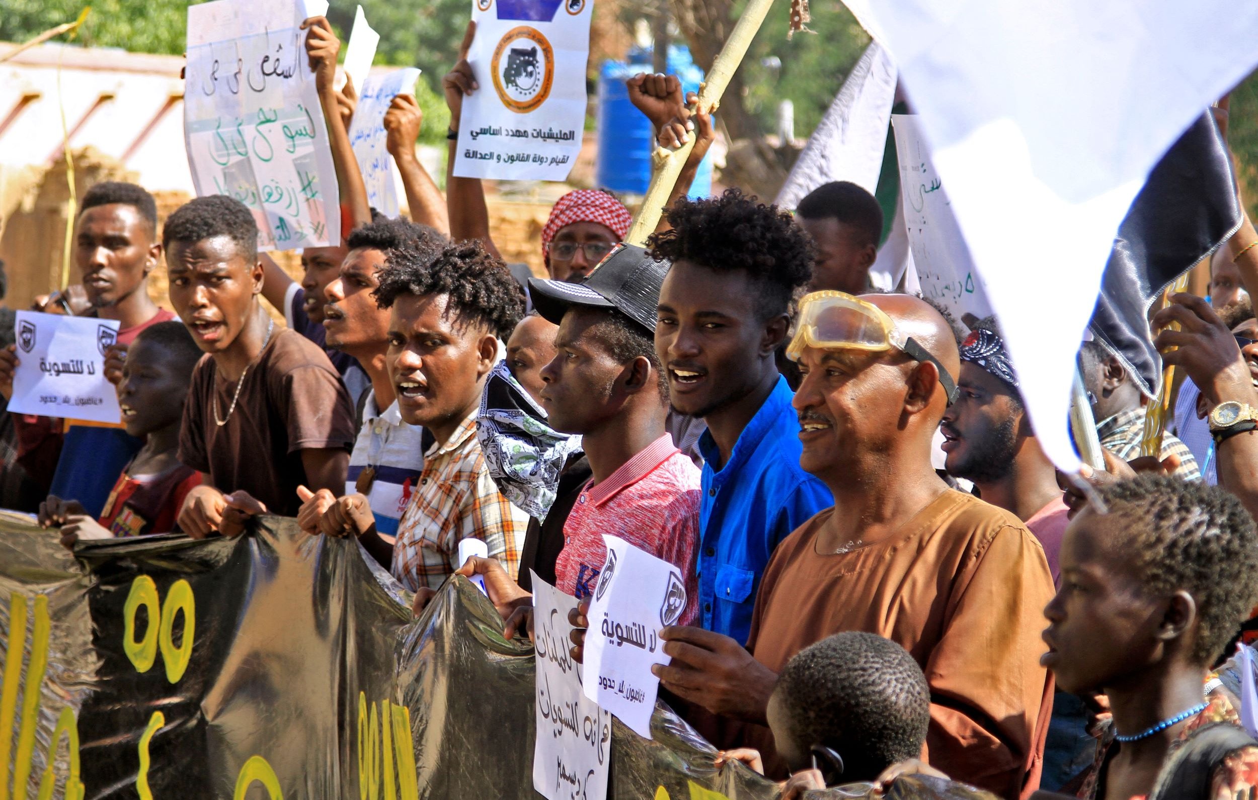 L'image montre un groupe de manifestants rassemblés, brandissant des pancartes et exprimant des slogans. Ils semblent engagés et passionnés, avec des expressions faciales intenses. Les gens portent des vêtements variés et il y a des drapeaux ou des bannières en arrière-plan. L'ambiance générale semble être celle d'une manifestation pour une cause précise.