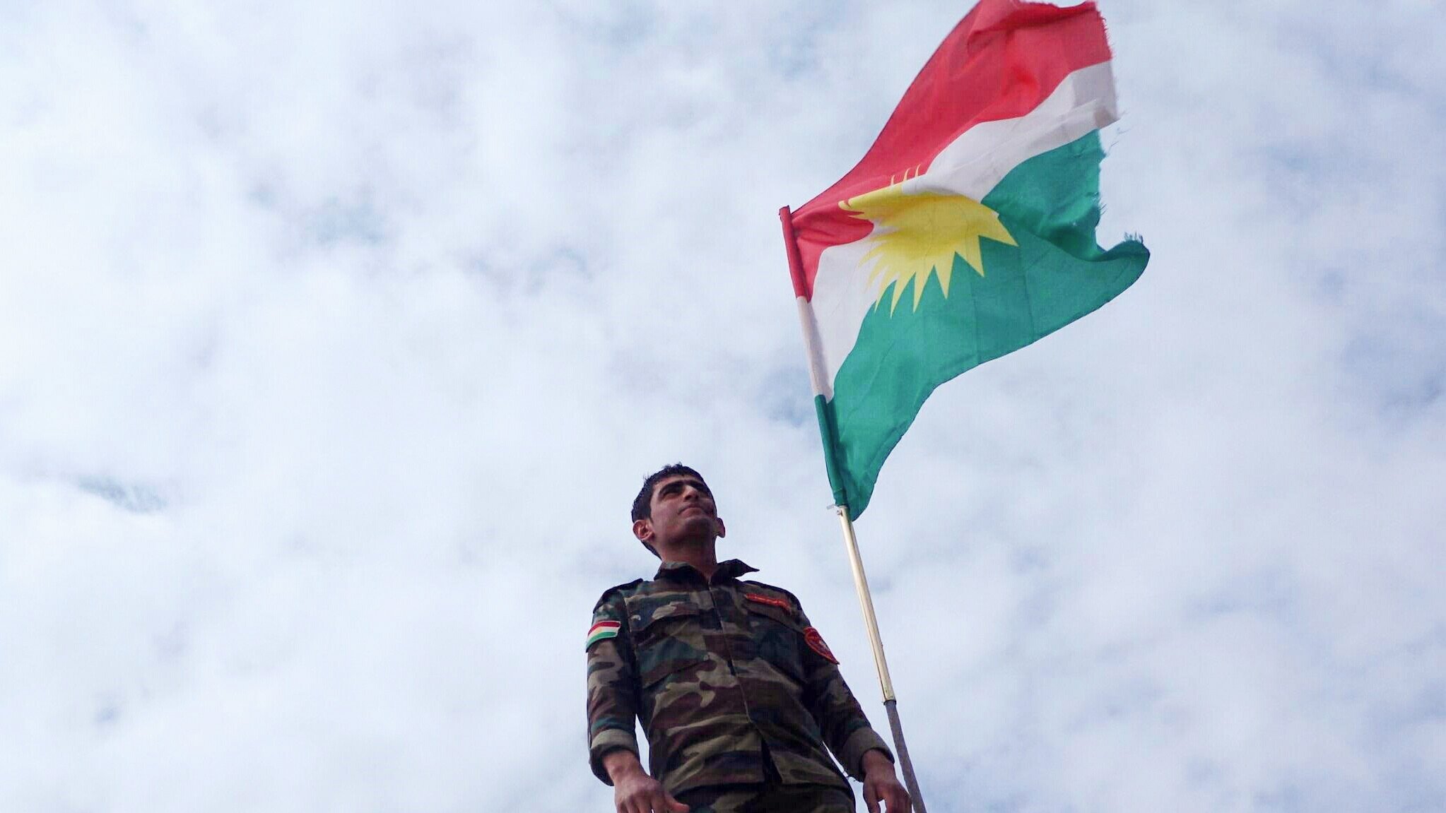 در تصویر، یک سرباز در کنار پرچم کردستان ایستاده است. او لباس نظامی به تن دارد و پرچم با رنگ‌های قرمز، سفید، سبز و دایره زرد رنگ را که نماد هویت کردی است، به اهتزار درآورده است. پس‌زمینه آسمان ابری است و این ترکیب به تصویر حس قوی و نمادین می‌دهد.