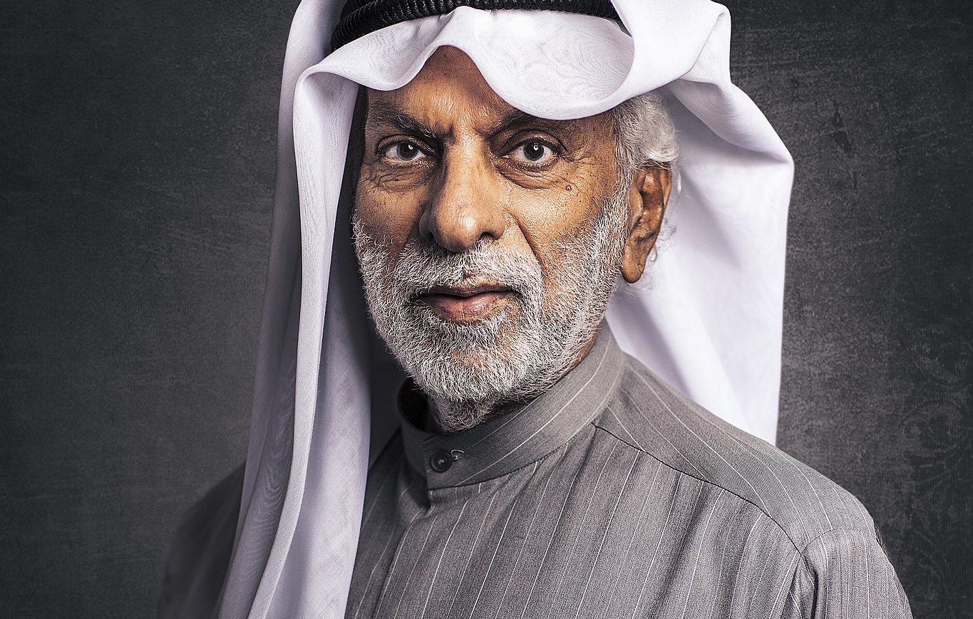 L'image montre un homme âgé avec une barbe grise, portant un vêtement traditionnel de style arabe, typiquement un thobe gris avec des rayures. Il a également un agal noir et un ghutrah blanche, qui est un couvre-chef traditionnel. L'arrière-plan est sombre, ce qui met en valeur son expression sage et son apparence traditionnelle.