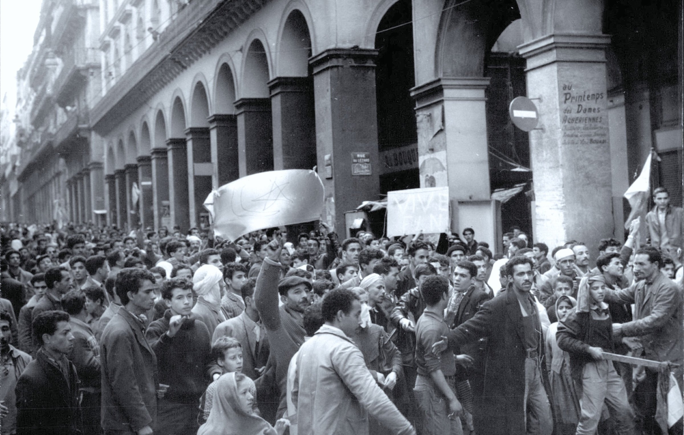 L'image montre une grande manifestation dans une rue animée, où une foule dense est rassemblée. Les manifestants semblent exprimer des revendications, brandissant des drapeaux et des pancartes. L'architecture environnante est typiquement urbaine, avec de grands arcs et des bâtiments anciens. L'atmosphère générale semble passionnée et engagée, reflétant un fort esprit de protestation.