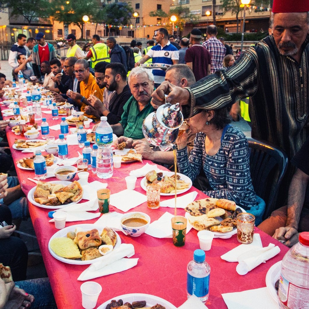 L'image montre une grande table dressée en plein air, où des personnes diverses sont réunies autour d'un repas convivial. Les convives, de différents âges, sont assis sur des chaises en plastique et profitent de divers plats, avec des assiettes remplies de nourriture. On peut voir des bouteilles d'eau sur la table et des verres. L'ambiance semble festive et chaleureuse, marquée par des sourires et des échanges entre les participants. En arrière-plan, on aperçoit d'autres personnes et des éléments de l'environnement urbain, indiquant une atmosphère de communauté.