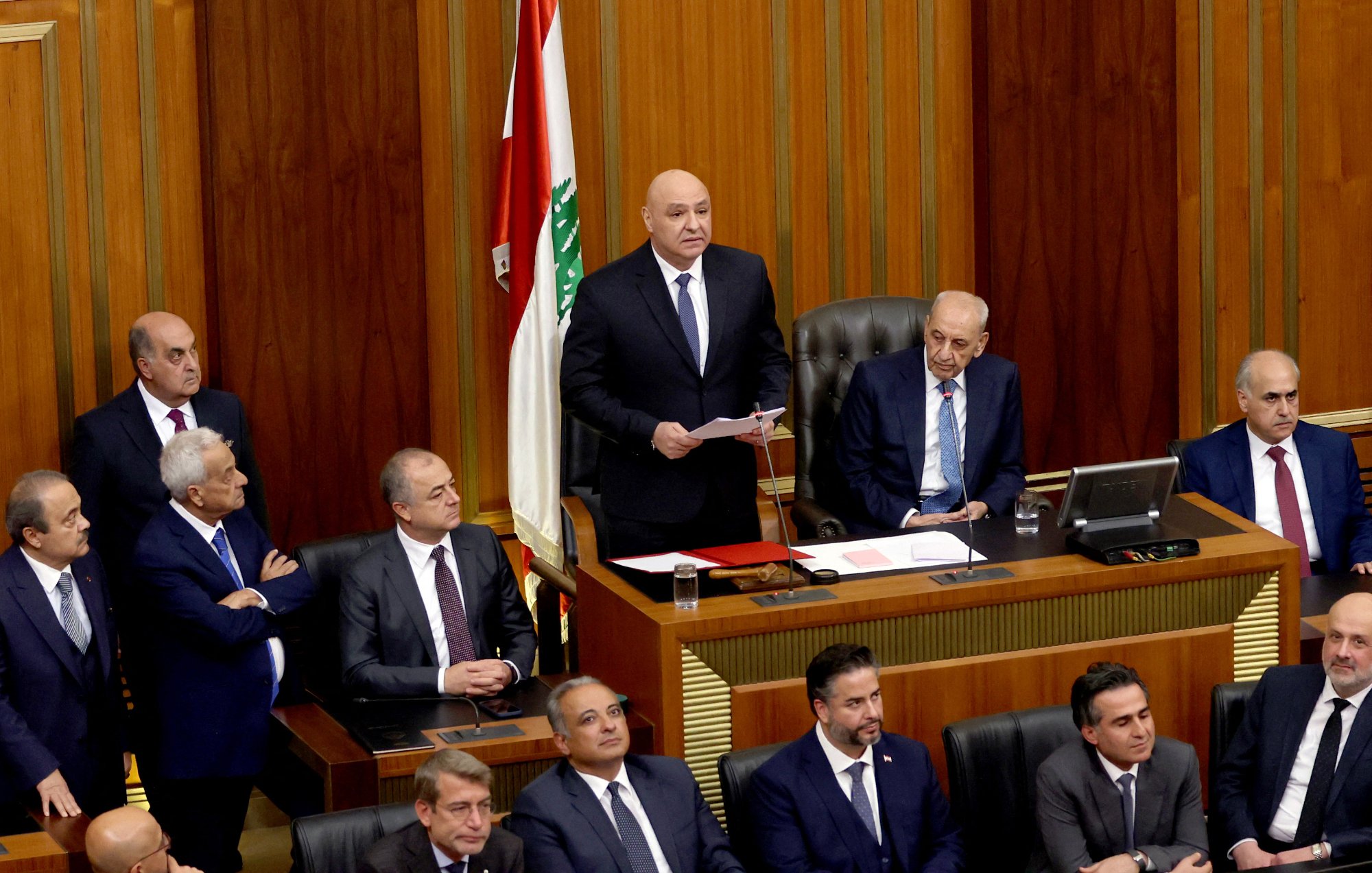 L'image présente une scène politique au sein d'un parlement. Au centre, un homme se tient debout, tenant des documents, probablement en train de s'adresser aux autres membres. À sa droite, un homme est assis derrière un bureau, semblant être un président ou un responsable de la séance. Plusieurs personnes sont debout et assises autour, portant des costumes, et affichent diverses expressions. En arrière-plan, on aperçoit le drapeau libanais. L'ambiance semble formelle et sérieuse, typique des assemblées législatives.