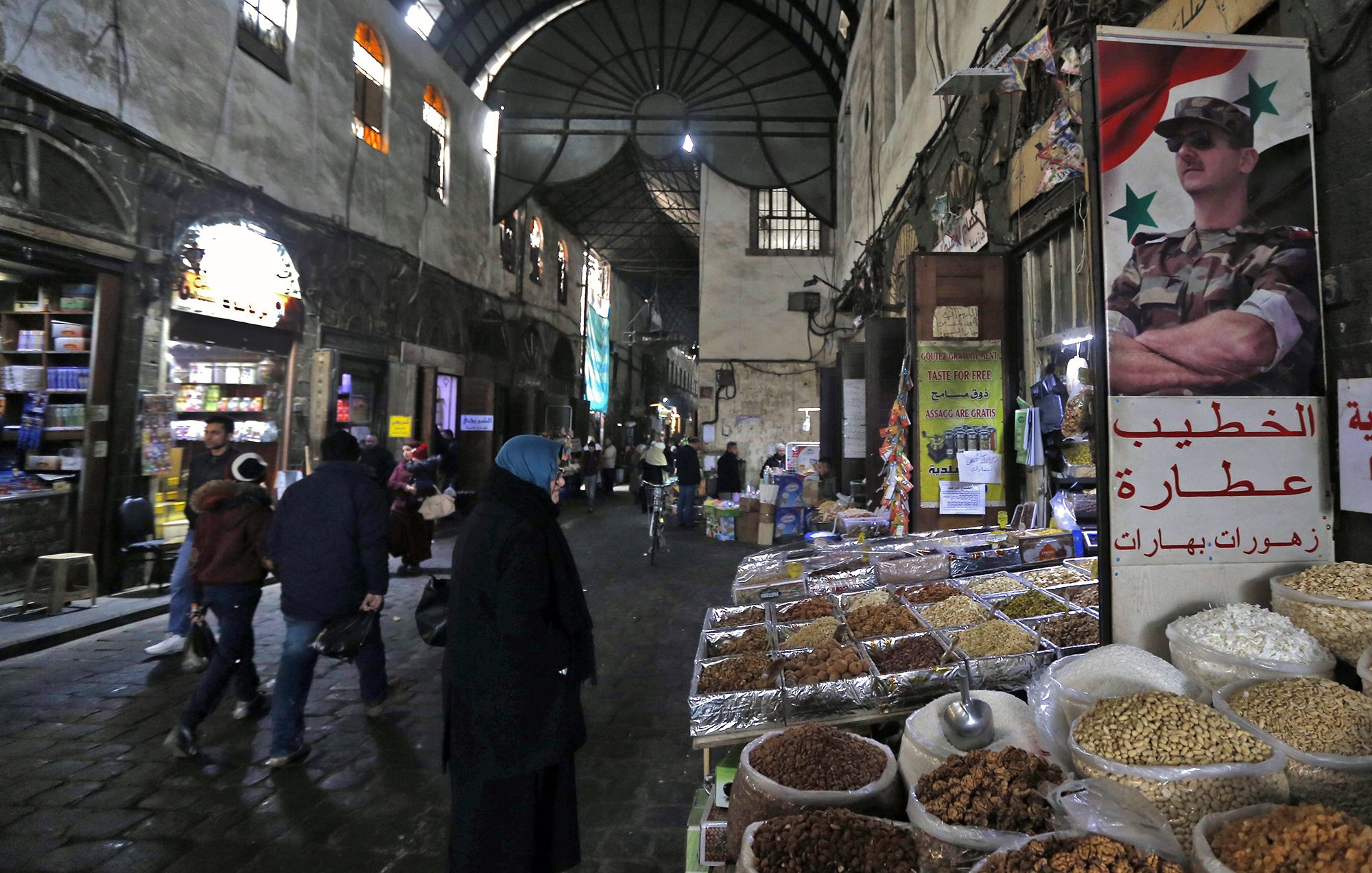 L'image montre une scène d'un souk ou d'un marché typique, probablement en Syrie. On aperçoit des étals avec des épices, des fruits secs, et d'autres produits en vente. Les murs de la rue sont en pierre et semblent anciens, créant une ambiance historique. Des personnes, dont certaines portent des vêtements traditionnels, se déplacent dans la rue, tandis qu'une affiche avec un symbole militaire est accrochée à un mur, ajoutant un élément de contexte culturel et politique à la scène. La lumière faible et les ombres accentuent l'ambiance conviviale et animée du marché.