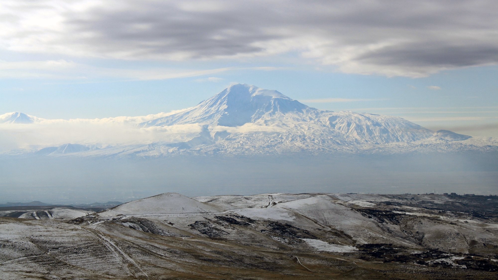 L'image montre un paysage montagneux impressionnant, avec un grand sommet enneigé qui semble être un volcan. Le sommet est entouré de nuages légers, créant une atmosphère mystique. Au premier plan, des collines aux teintes terreuses et parfois enneigées s'étendent, ajoutant à la beauté naturelle de la scène. Le ciel est partiellement couvert, laissant transparaître une lumière douce qui éclaire le paysage.