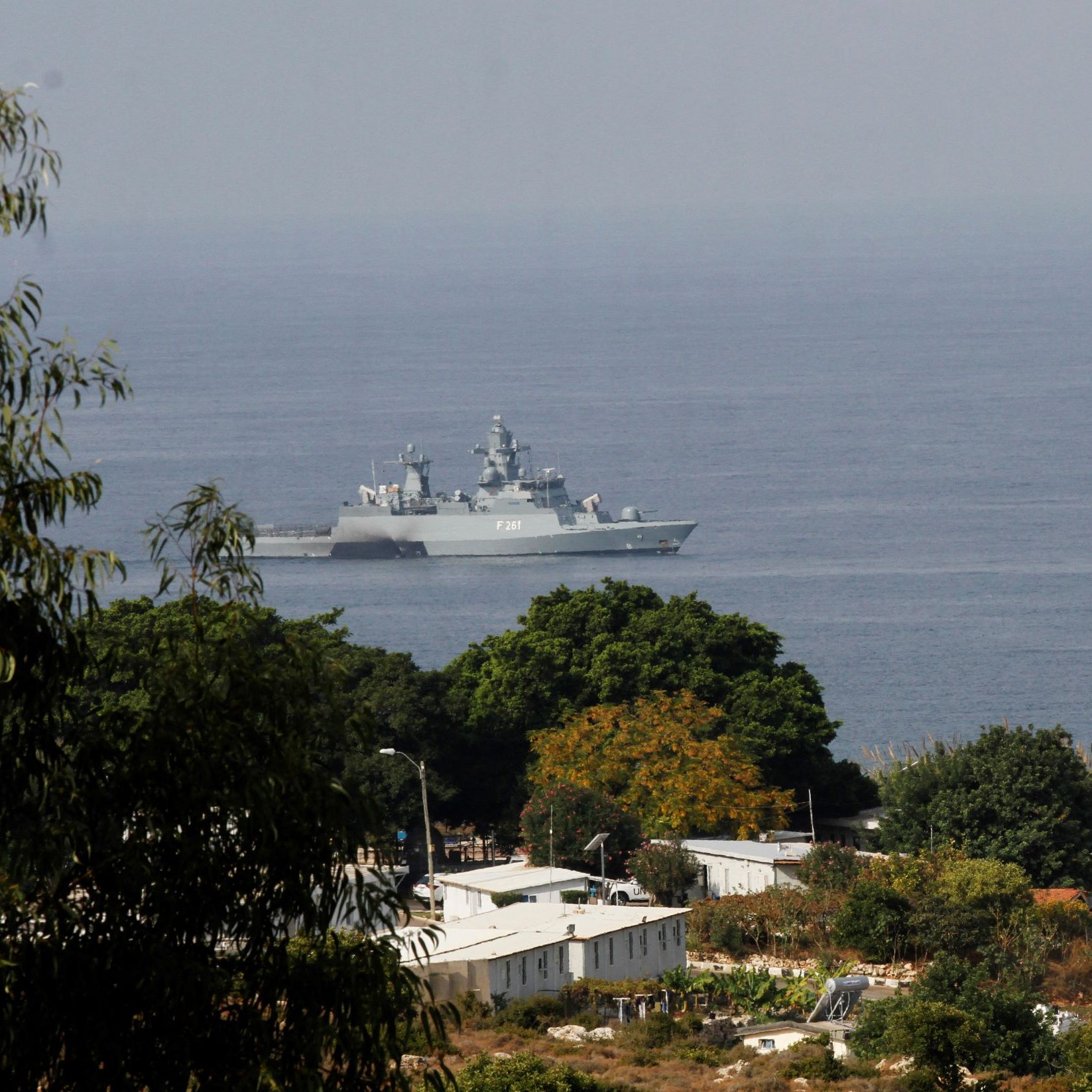 تظهر في الصورة سفينة حربية تجوب المياه بالقرب من ساحل يطل على منطقة خضراء. في الخلفية، يمكن رؤية بعض المباني والمرافق، كما يُلاحظ وجود برج مراقبة. السماء تبدو صافية، والمشهد يعكس أجواء بحرية هادئة.
