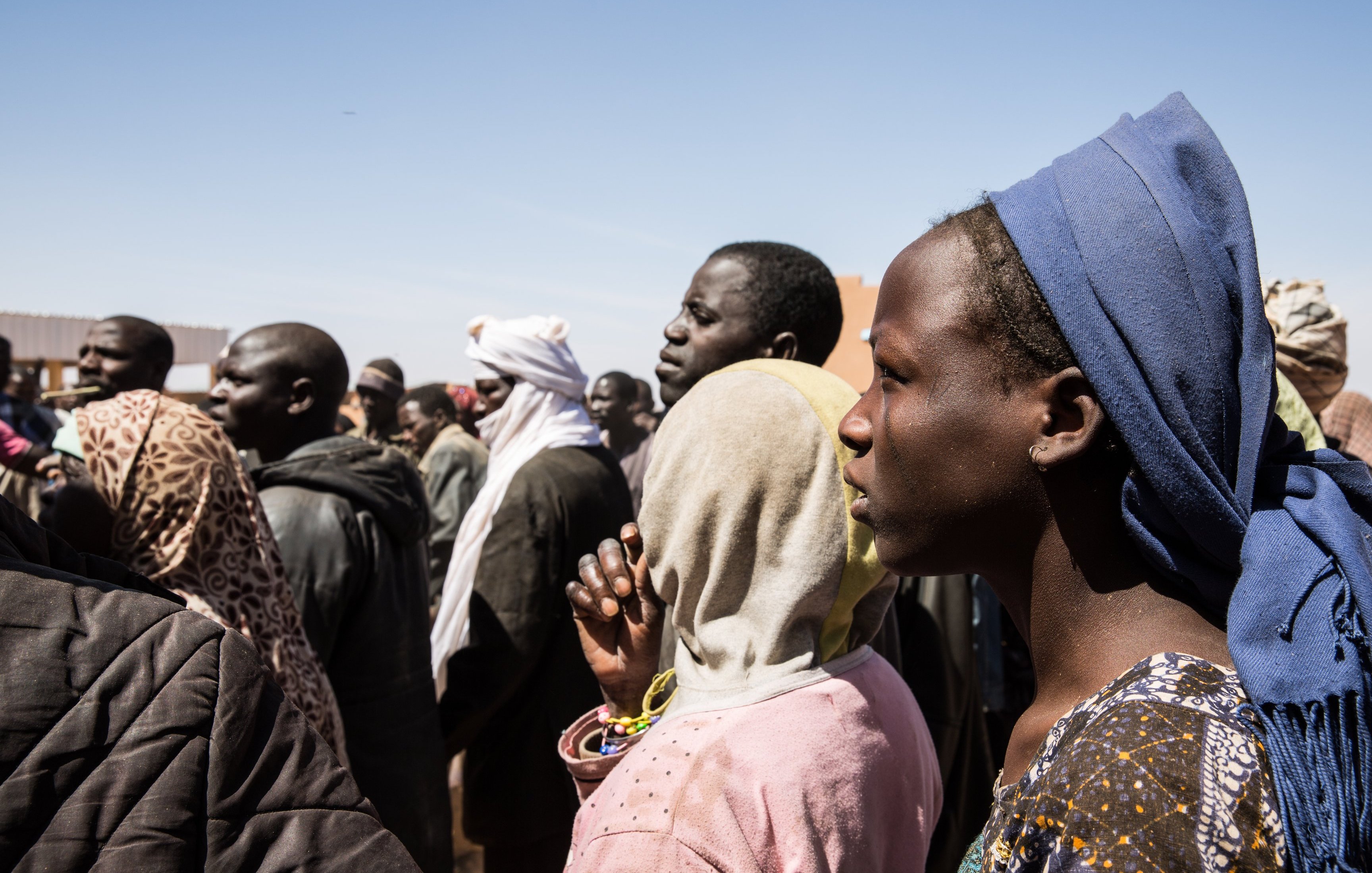 L'image montre un groupe de personnes, probablement en attente ou rassemblées pour une raison quelconque. On peut voir des visages divers avec des expressions sérieuses. Certaines personnes portent des vêtements traditionnels et des accessoires variés, comme des écharpes ou des foulards. L'arrière-plan est flou, mais le ciel est dégagé. L'ambiance semble concentrée, peut-être en lien avec un événement important.