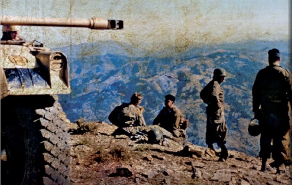 L'image montre un groupe de soldats situés sur un sommet de montagne. À gauche, un char de combat est visible, sa tourelle tournée vers l'avant. Les soldats sont en uniformes militaires, certains semblent assis ou se tiennent debout, avec des paysages montagneux s'étendant en arrière-plan. L'environnement paraît aride, et la lumière donne une atmosphère de calme malgré la présence militaire. Les couleurs de l'image évoquent une certaine nostalgie, suggérant qu'elle pourrait provenir d'une période passée, peut-être de la guerre.