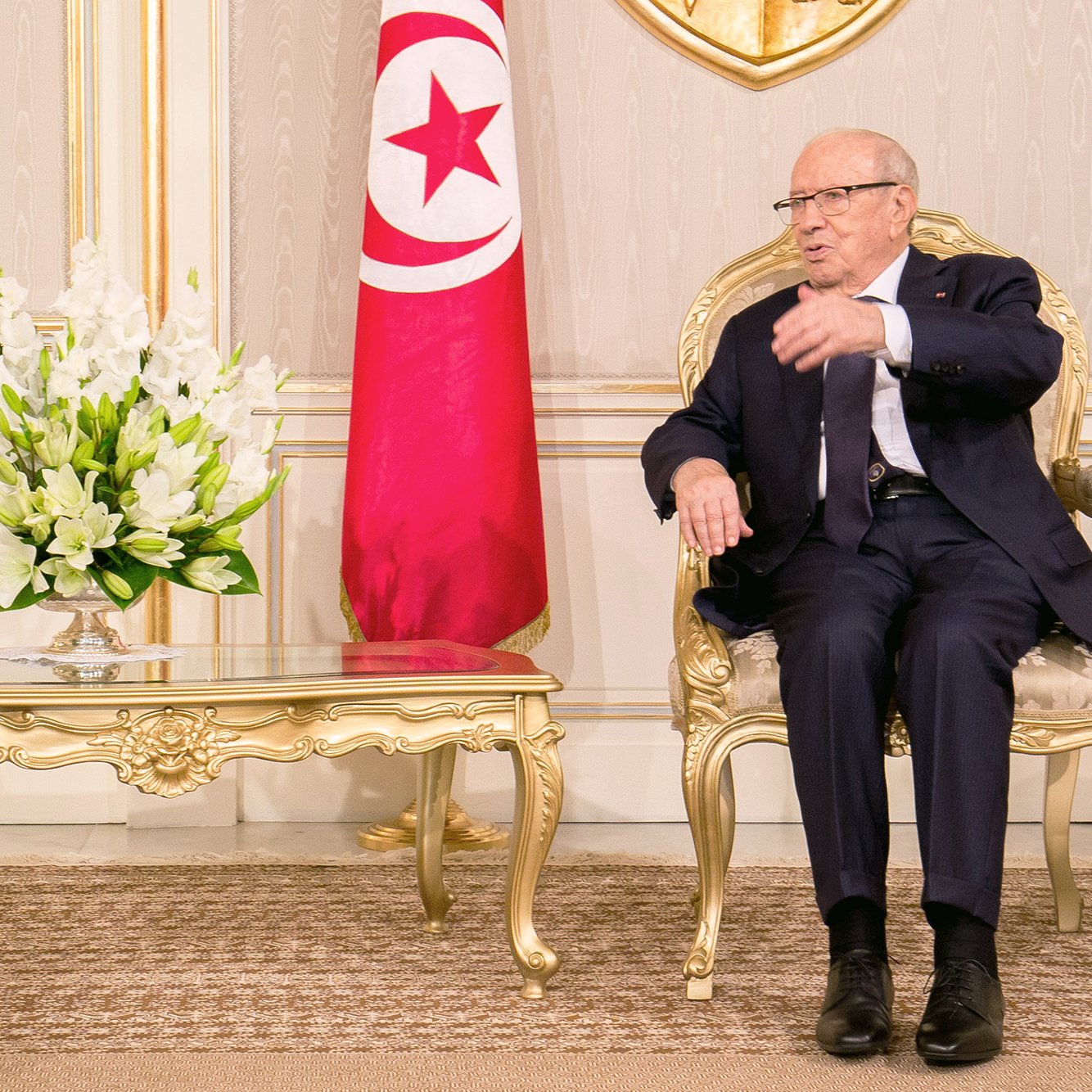 الصورة تظهر رجلًا يجلس على كرسي فاخر في غرفة مزينة بشكل أنيق. خلفه علم تونس، وهناك طاولة صغيرة أمامه تحتوي على زهور بيضاء. يبدو الرجل في وضع يتحدث أو يشرح شيئًا، ويرتدي بدلة رسمية وقميص. الغرفة تبرز بديكوراتها المثالية وألوانها الهادئة.