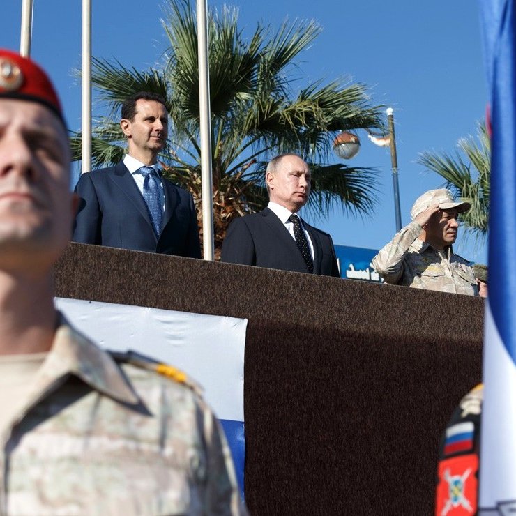 L'image montre un groupe de personnes, dont deux figures de haut rang, sur une plateforme en extérieur, avec un fond de palmiers et un ciel bleu. Au premier plan, des soldats en uniforme, probablement d'une unité militaire, sont présents, portant des drapeaux. Les expressions des personnes sur la plateforme semblent sérieuses et concentrées, indiquant un événement officiel ou militaire.