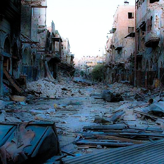 L'image montre une rue dévastée, probablement après un conflit ou une catastrophe. On peut voir des bâtiments endommagés avec des murs effondrés et des débris éparpillés sur le sol. Le contexte semble être celui d'une ville abandonnée, avec une atmosphère sombre et désolée. Les traces de destruction sont évidentes, illustrant les ravages subis par l'environnement urbain.