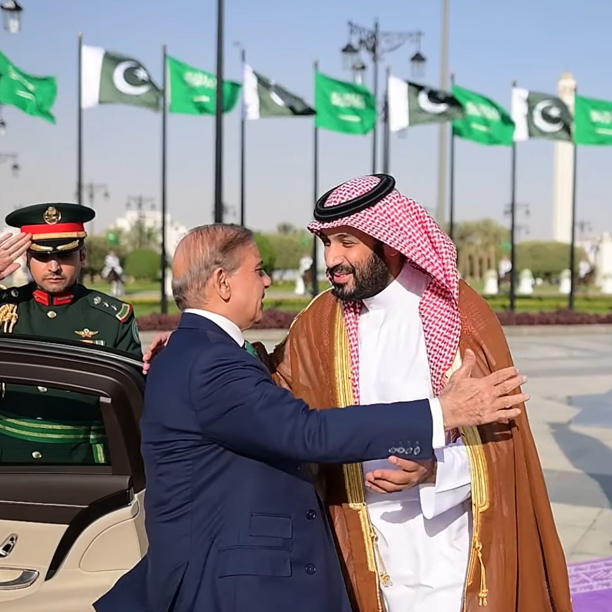 Deux hommes en costume échangent une accolade, avec des drapeaux en arrière-plan.