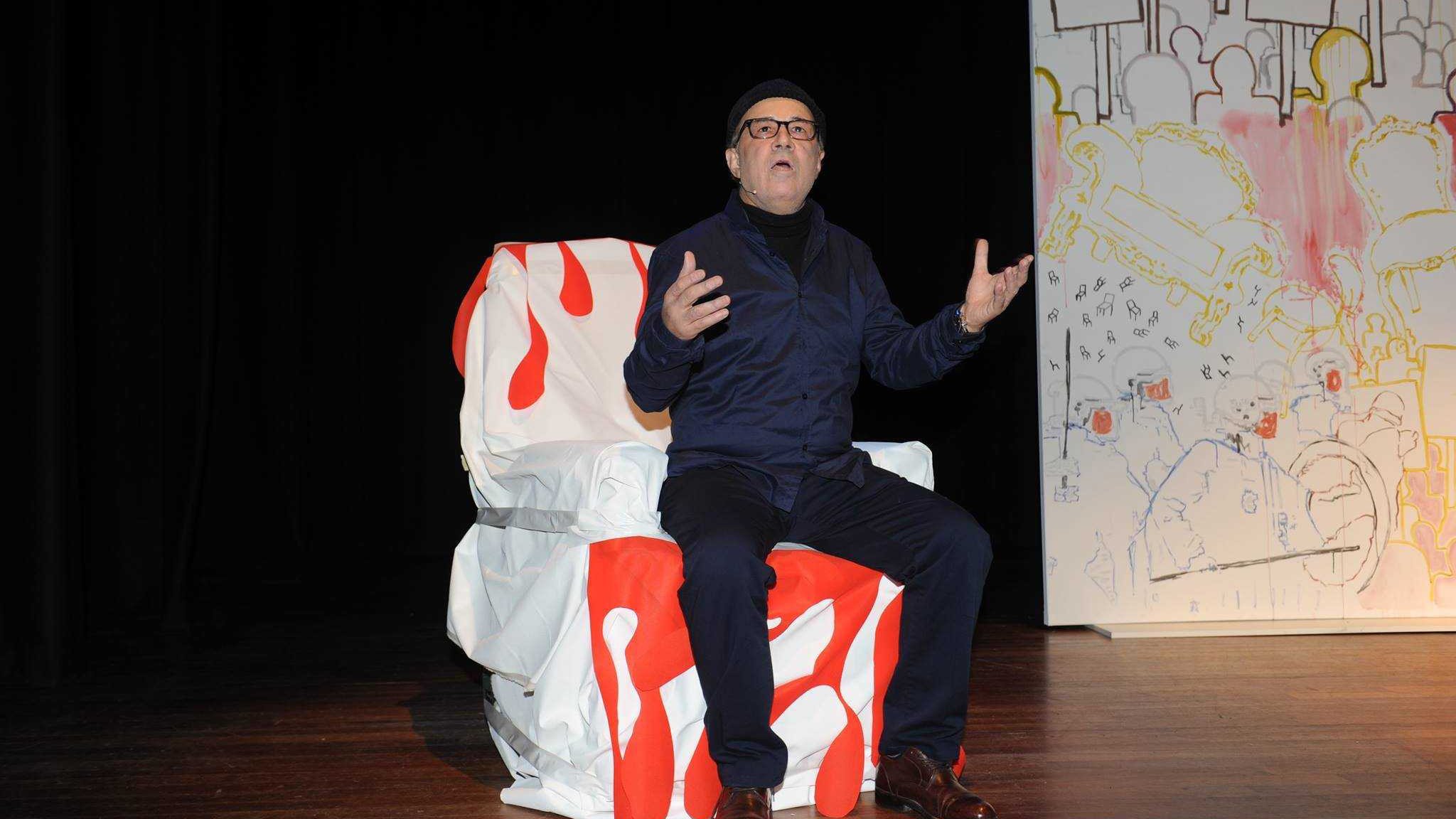 L'image montre un homme assis sur une chaise décorée de motifs rouges et blancs. Il est vêtu de vêtements sombres et porte un bonnet. L'homme a l'air expressif, gesticulant avec les mains, peut-être en train de parler ou de performer. En arrière-plan, on voit un panneau avec des dessins colorés et des formes abstraites. L'atmosphère semble être celle d'une scène de théâtre.