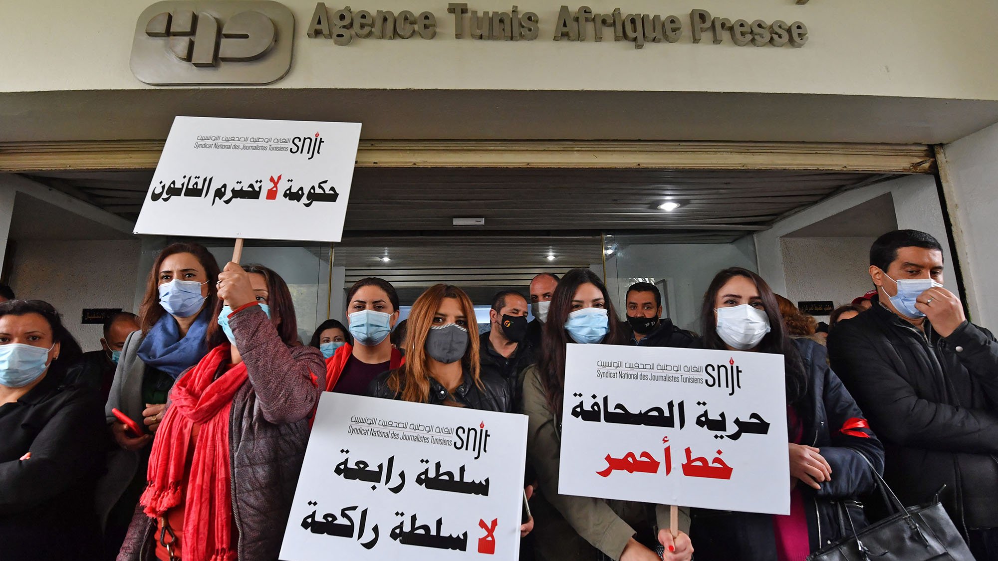 تظهر الصورة مجموعة من الأشخاص يقفون أمام مقر وكالة تونس أفريقيا للأنباء. يحملون لافتات تعبر عن مطالبهم، مثل "حكومة تخرّق القانون" و"حرية الصحافة خط أحمر". كما يبدو أنهم يضعون أقنعة للوجه، مما يشير إلى أنهم قد يكونون في حالة احتجاج أو تظاهرة تتعلق بحرية الصحافة. تبرز تعبيرات الجدية والعزم على وجوه المشاركين في الوقفة.