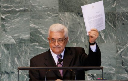 L'image montre un homme debout devant un pupitre de l'Organisation des Nations Unies (ONU). Il porte un costume noir avec une cravate et des lunettes. L'homme soulève un document, probablement un discours ou une déclaration, en s'adressant à l'assemblée. L'arrière-plan est décoré de panneaux en pierre, typiques des salles de conférence de l'ONU.