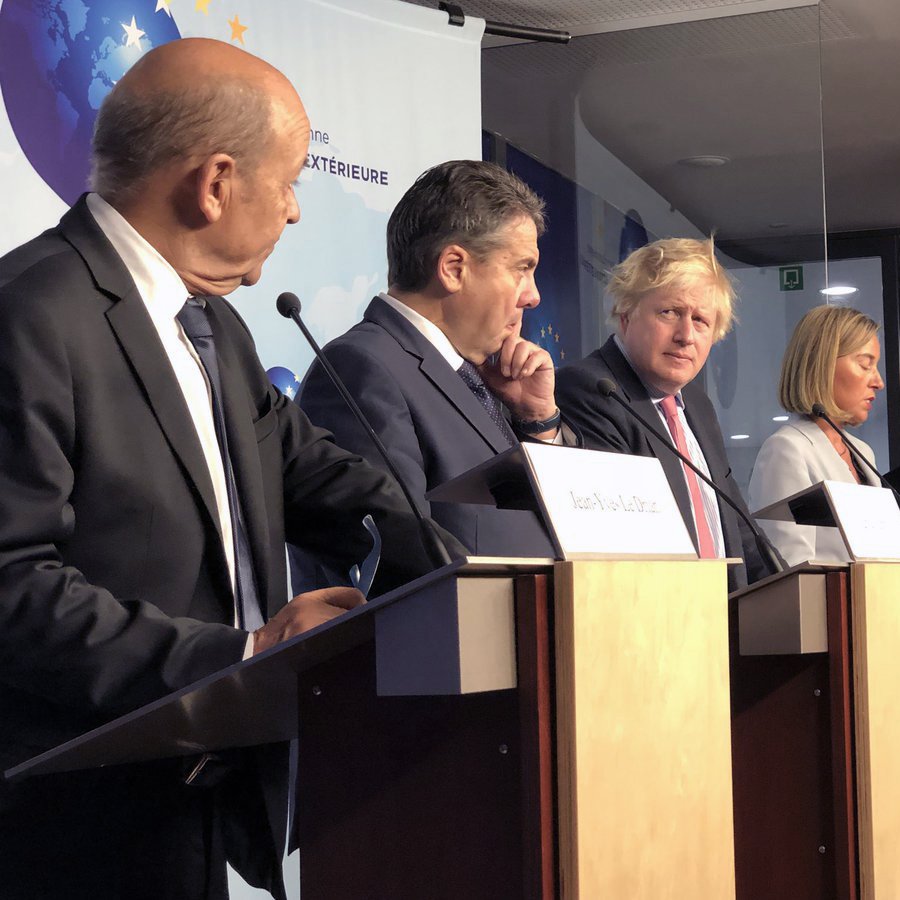 در این تصویر، گروهی از سیاستمداران در حال برگزاری یک کنفرانس خبری یا نشست مطبوعاتی هستند. چندین نفر بر روی چهار پایه ایستاده‌اند که هر کدام به یک میکروفن مجهز هستند. در میان آن‌ها مردی با موهای بلوند و کوتاه به چشم می‌خورد که به نظر می‌رسد یکی از افراد کلیدی این نشست است. سایر افراد نیز در حالت‌های مختلف ایستاده و به صحبت‌های یکدیگر گوش می‌دهند. پس‌زمینه تصویر شامل یک صفحه نمایش یا دیوار با نشانه‌های مربوط به جلسه است. این تصویر به فضای رسمی و دیپلماتیک اشاره دارد.