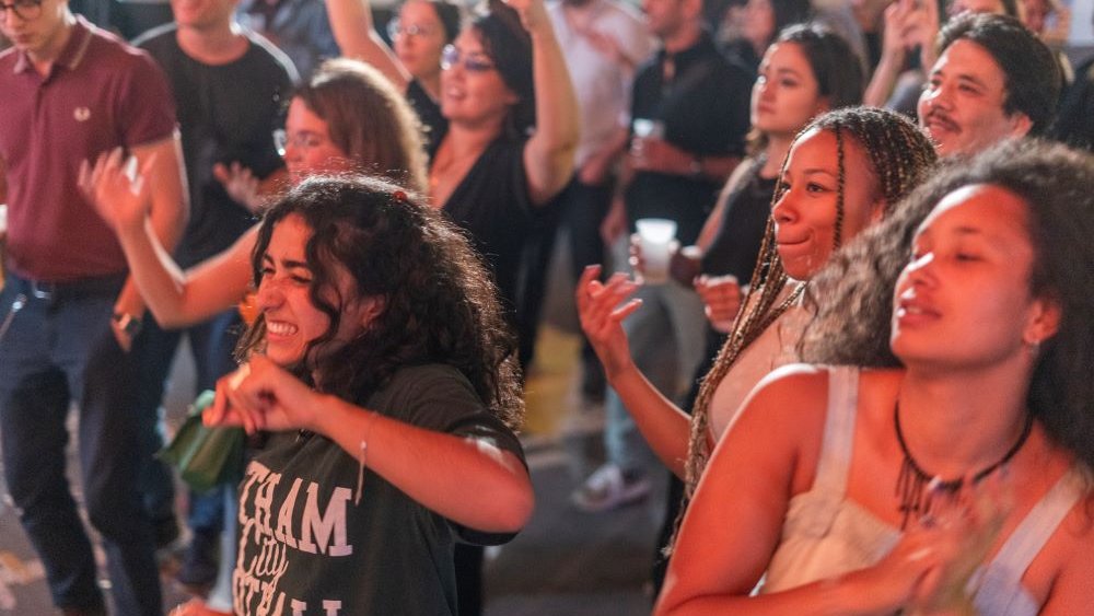 L'image montre un groupe de personnes en train de danser et de profiter d'une ambiance festive. On peut voir des personnes de différents âges, souriantes et s'amusant. Certaines gesticulent au rythme de la musique, tandis que d'autres sont concentrées sur le moment. L'éclairage est chaleureux, ce qui contribue à l'atmosphère joyeuse de l'événement.