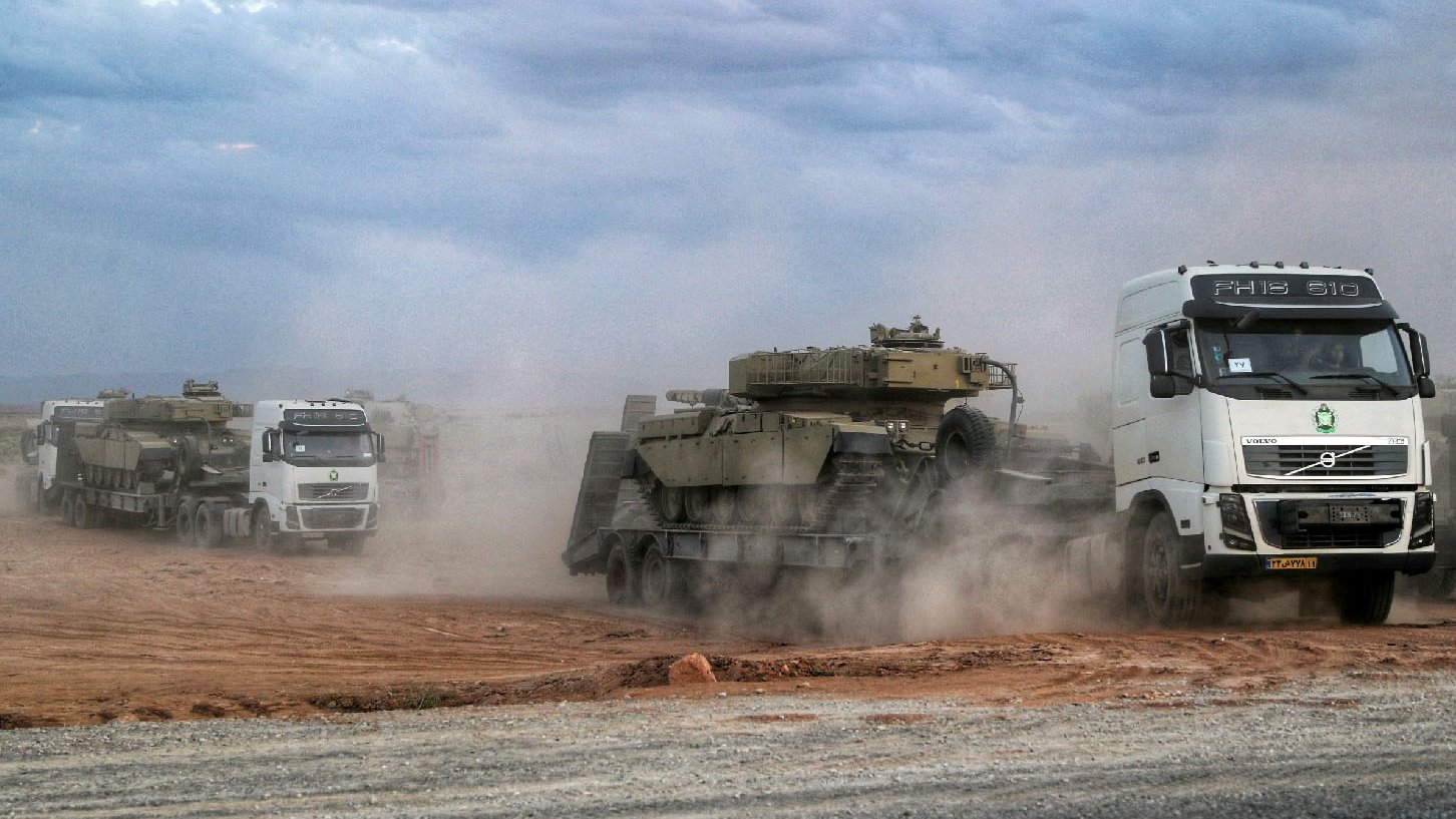 L'image montre une scène de transport militaire, avec plusieurs camions, notamment un portant le logo Volvo, transportant des véhicules blindés. La poussière s'élève autour des véhicules, suggérant un environnement saharien ou aride. Le ciel est nuageux, ajoutant à l'atmosphère de la scène. Les camions avancent sur une route en terre, mettant en avant un aspect logistique lié aux opérations militaires.