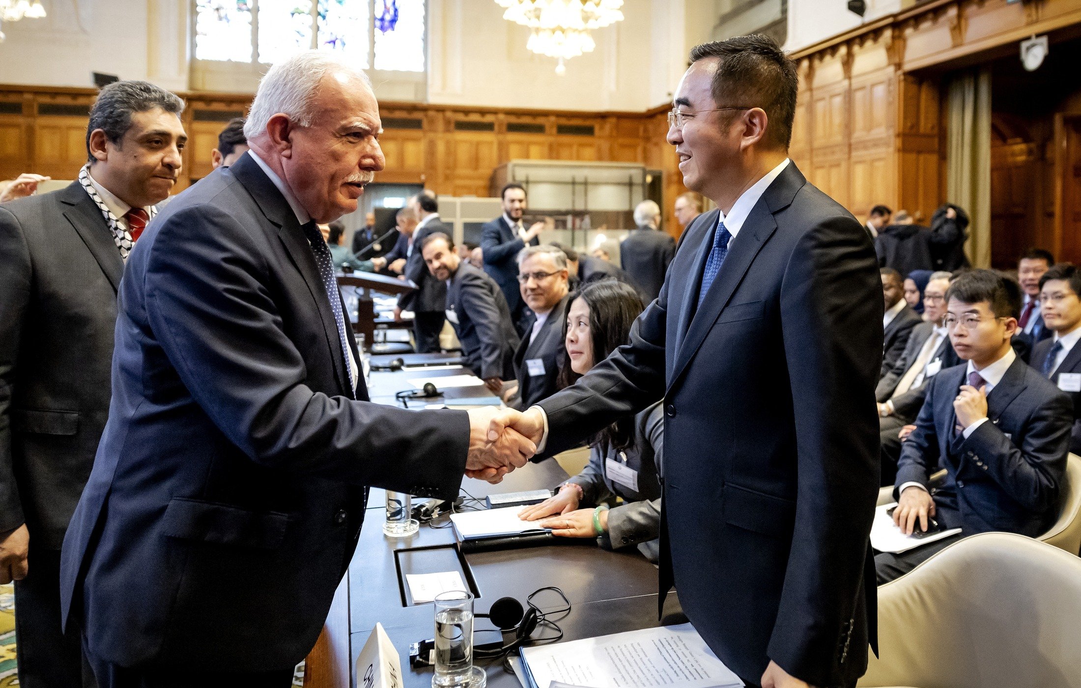 La imagen muestra a un grupo de personas en un entorno de reunión, probablemente en una conferencia o cumbre. En primer plano, dos hombres se están estrechando la mano, lo que sugiere un saludo cordial o un acuerdo. Uno de ellos tiene el cabello canoso y viste un traje oscuro, mientras que el otro es más joven y usa un traje también oscuro. Al fondo, se pueden ver otras personas sentadas alrededor de una mesa, con documentos y laptops, observando la interacción. El ambiente es formal y parece que se trata de un encuentro importante.